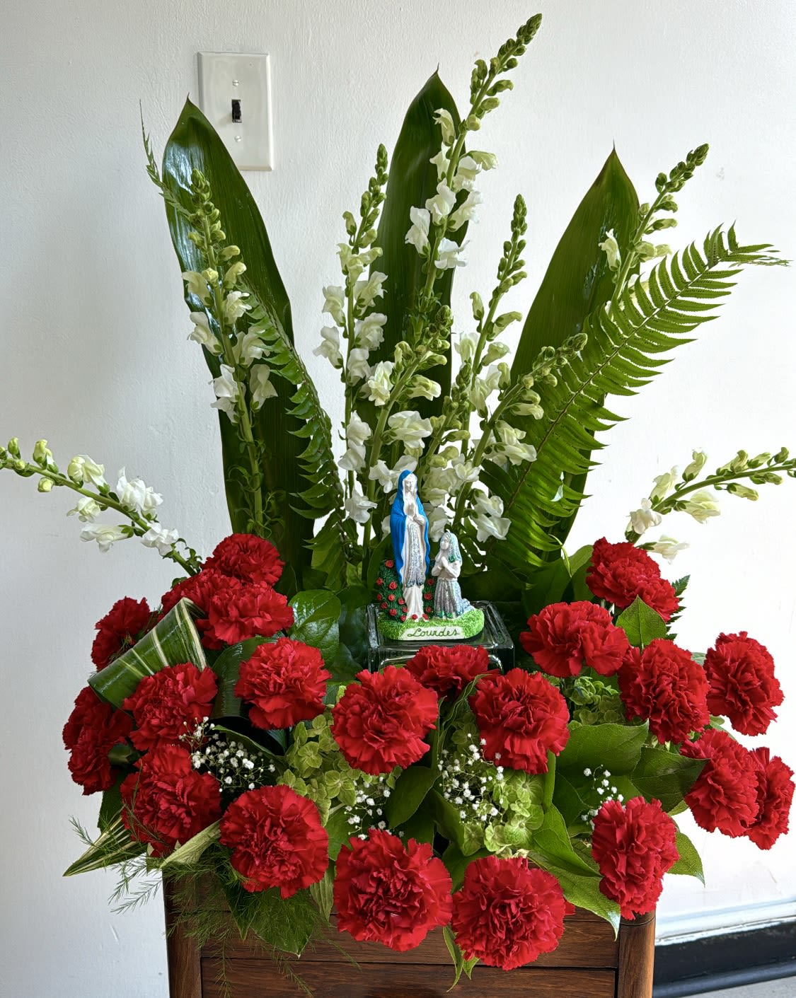 Red carnations with white flowers and a small Lourdes statue in a wooden container