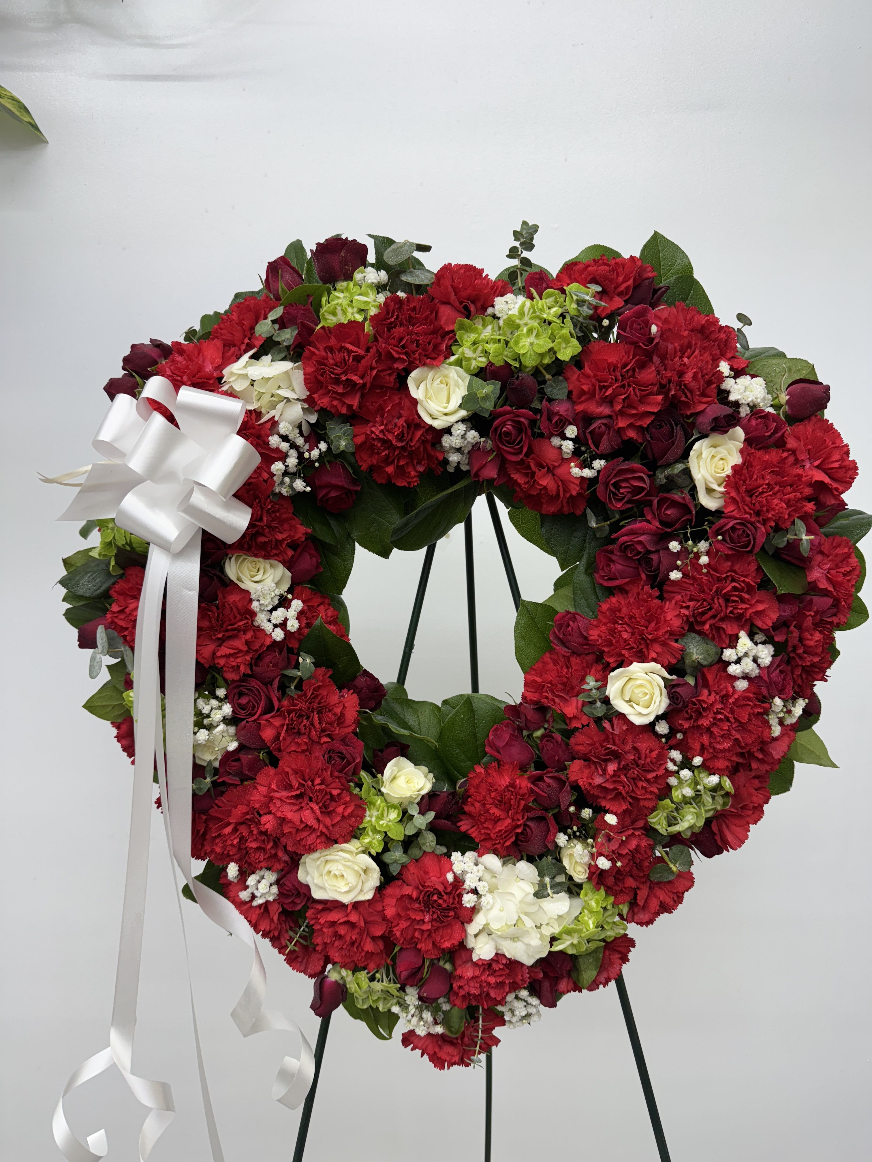 Heart-shaped floral wreath with red carnations and roses, accented by a white ribbon bow