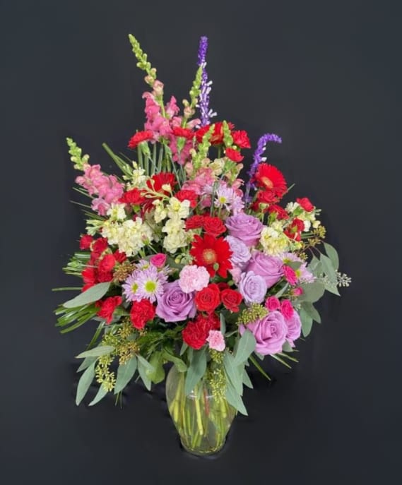 Tall mixed arrangement of red, pink, yellow, and lavender flowers in a clear glass vase