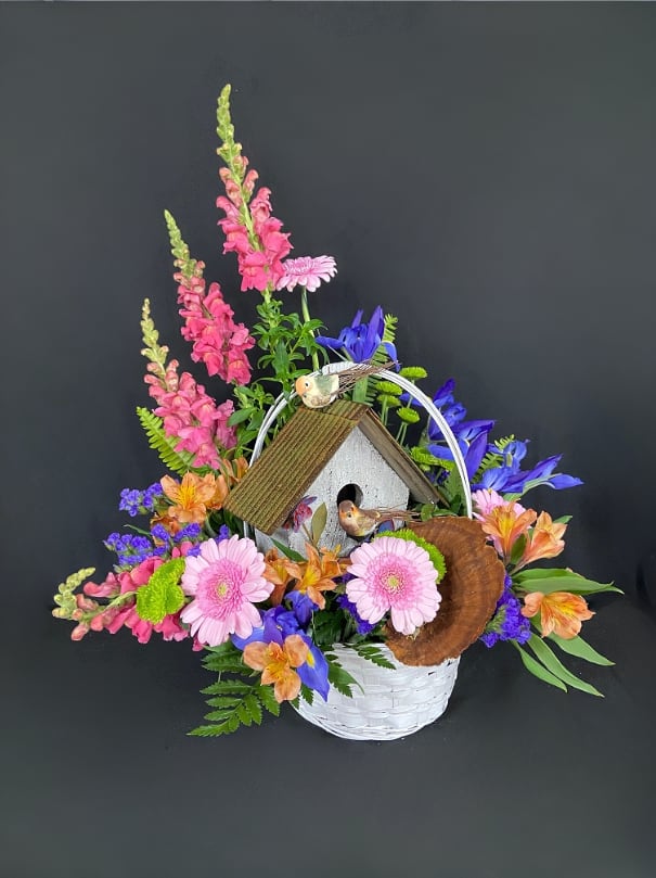 Basket arrangement with pink snapdragons, gerbera daisies, blue irises and a birdhouse centerpiece
