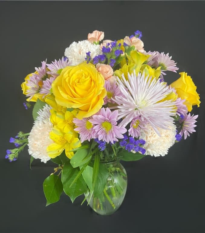 Mixed bouquet of yellow roses, lavender daisies, and white blooms in a clear glass vase