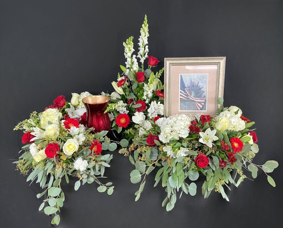 Patriotic red and white floral display with urn and framed flag photo against a black background