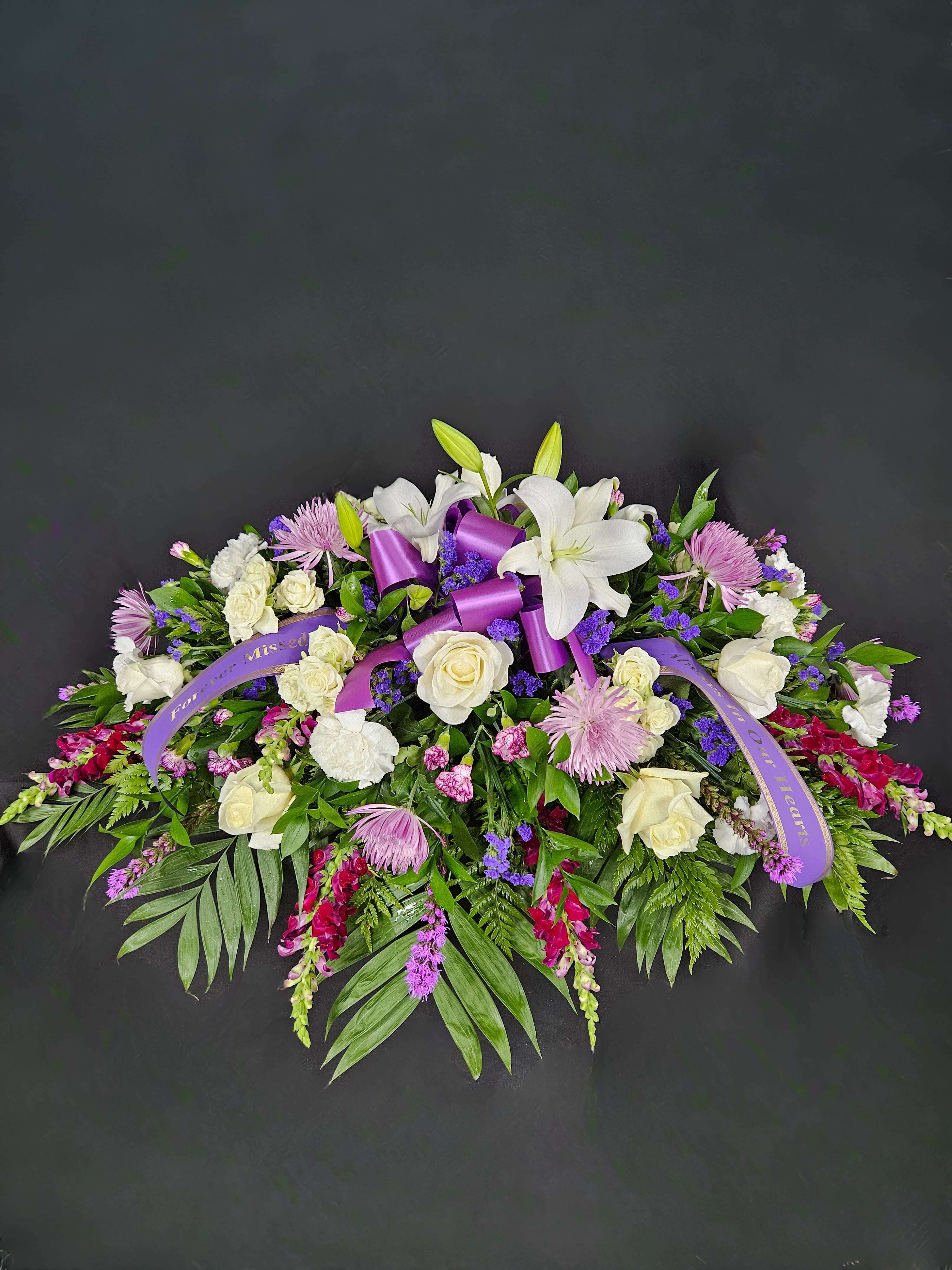 Elongated spray of white lilies, roses and purple flowers with a purple sympathy ribbon on a dark background