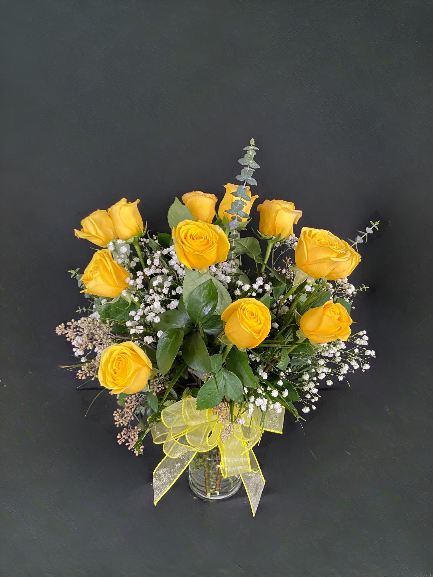 Arrangement of yellow roses with white filler flowers in a clear glass vase with a yellow bow.