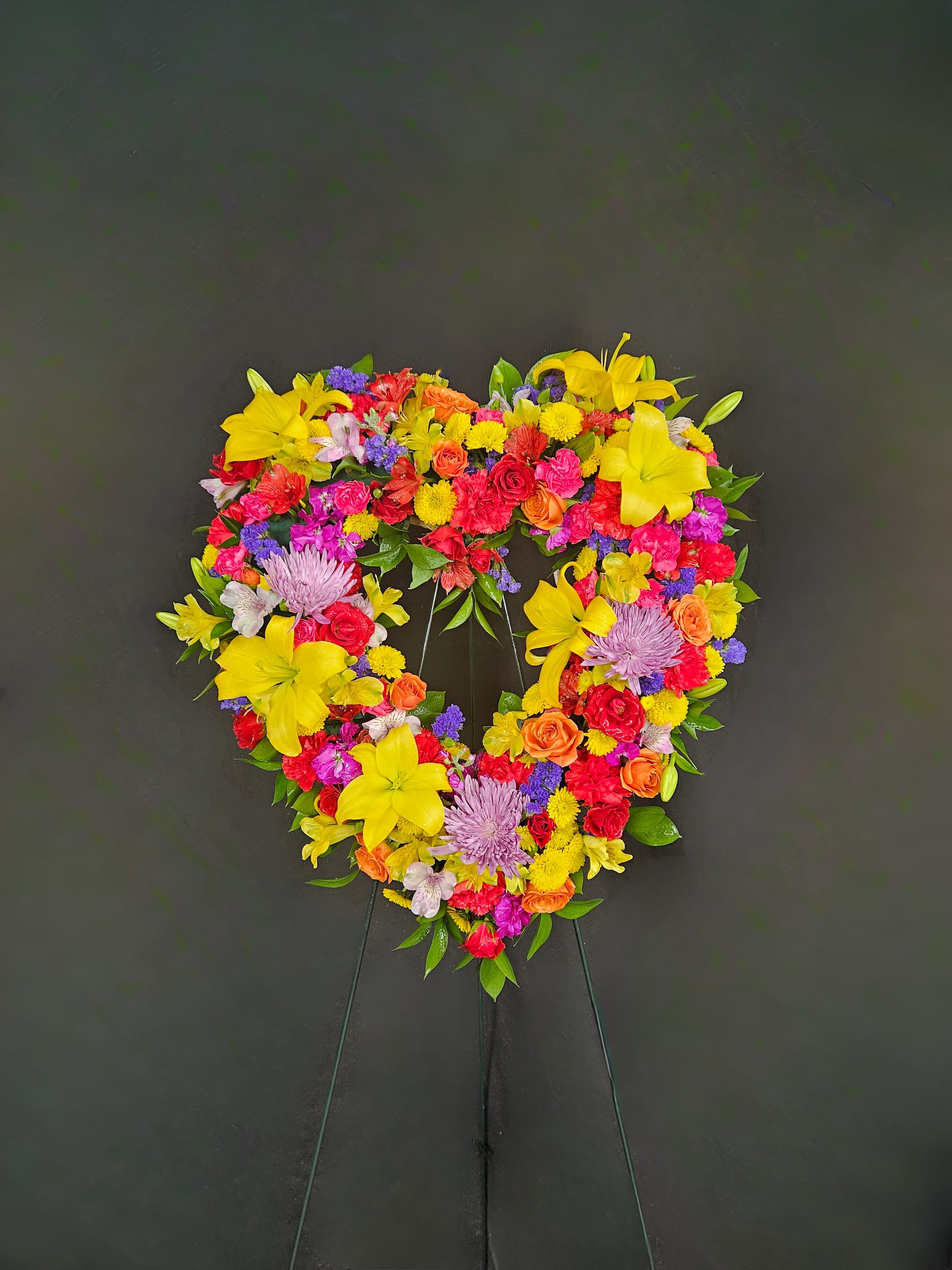 Heart-shaped standing wreath of bright yellow lilies, roses, carnations, and mums on a dark background