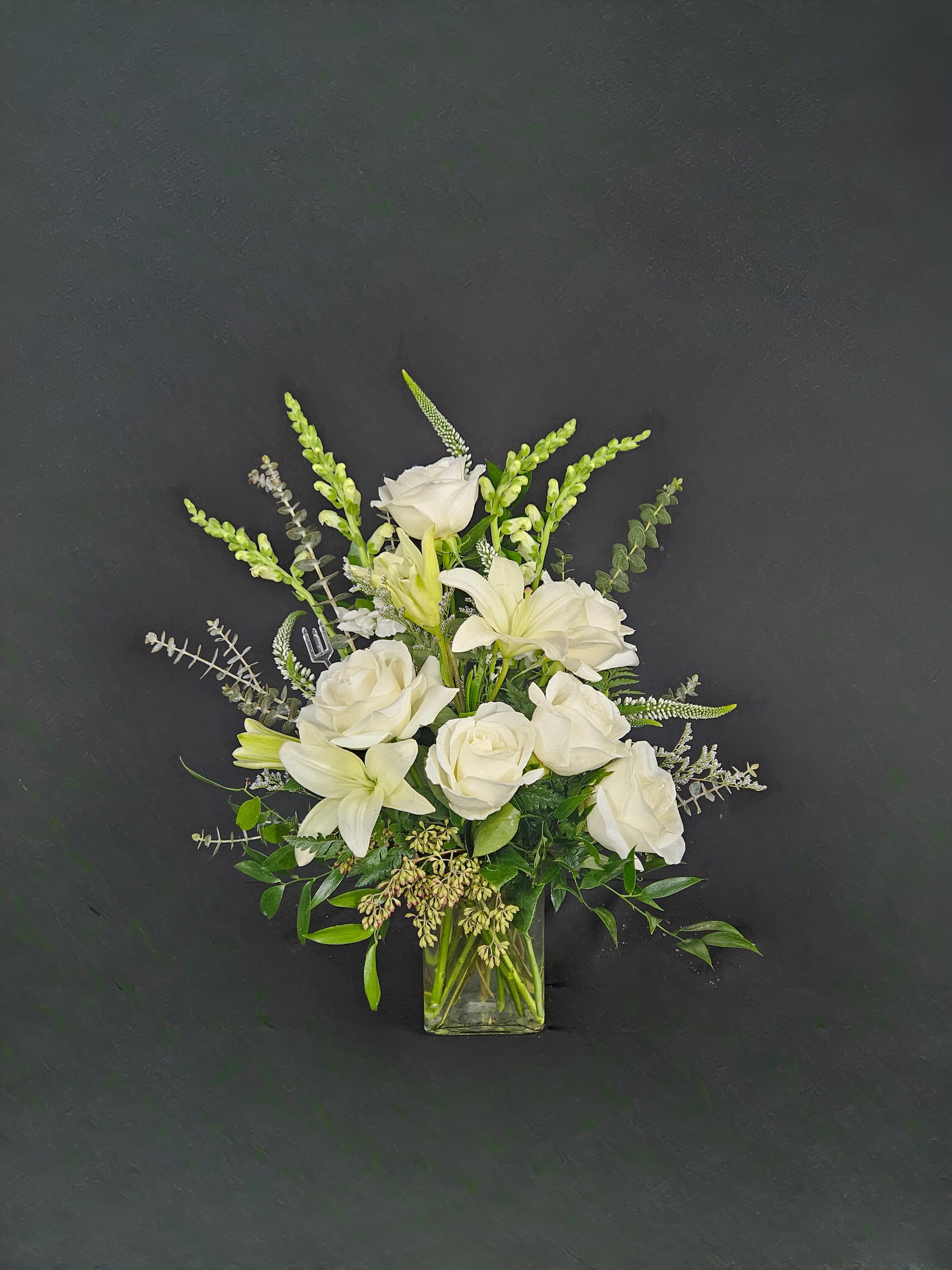 White roses, lilies, and snapdragons arranged in a clear glass vase against a dark background