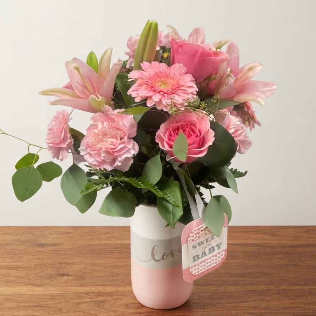 Pink mixed bouquet in a white and pink vase with a baby tag
