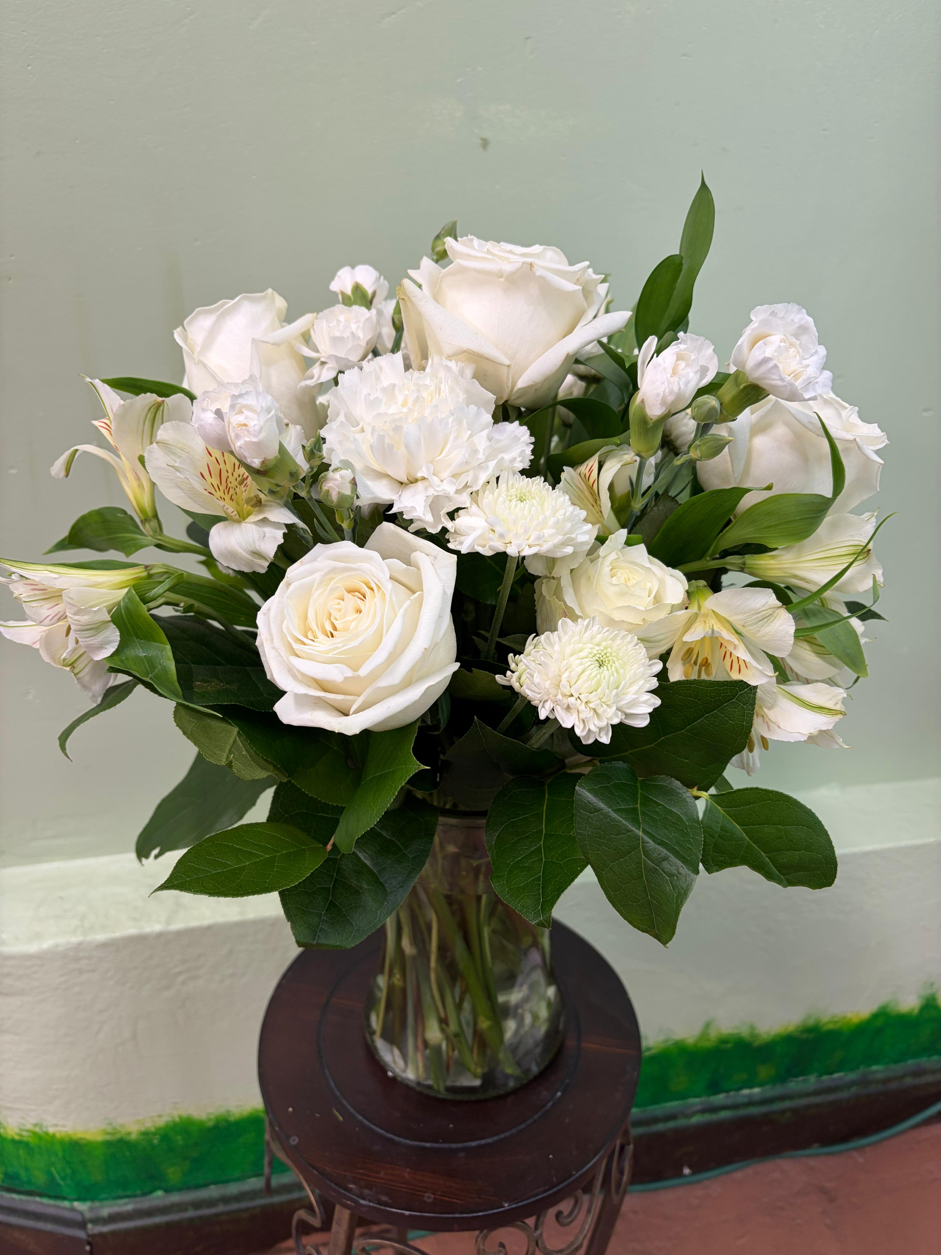 Ivory Grace - A timeless arrangement featuring creamy white roses, soft carnations, and delicate alstroemeria, accented with lush greenery. Designed in a clear glass vase, this piece brings a sense of calm, purity, and natural beauty—perfect for any occasion from sympathy to celebration.