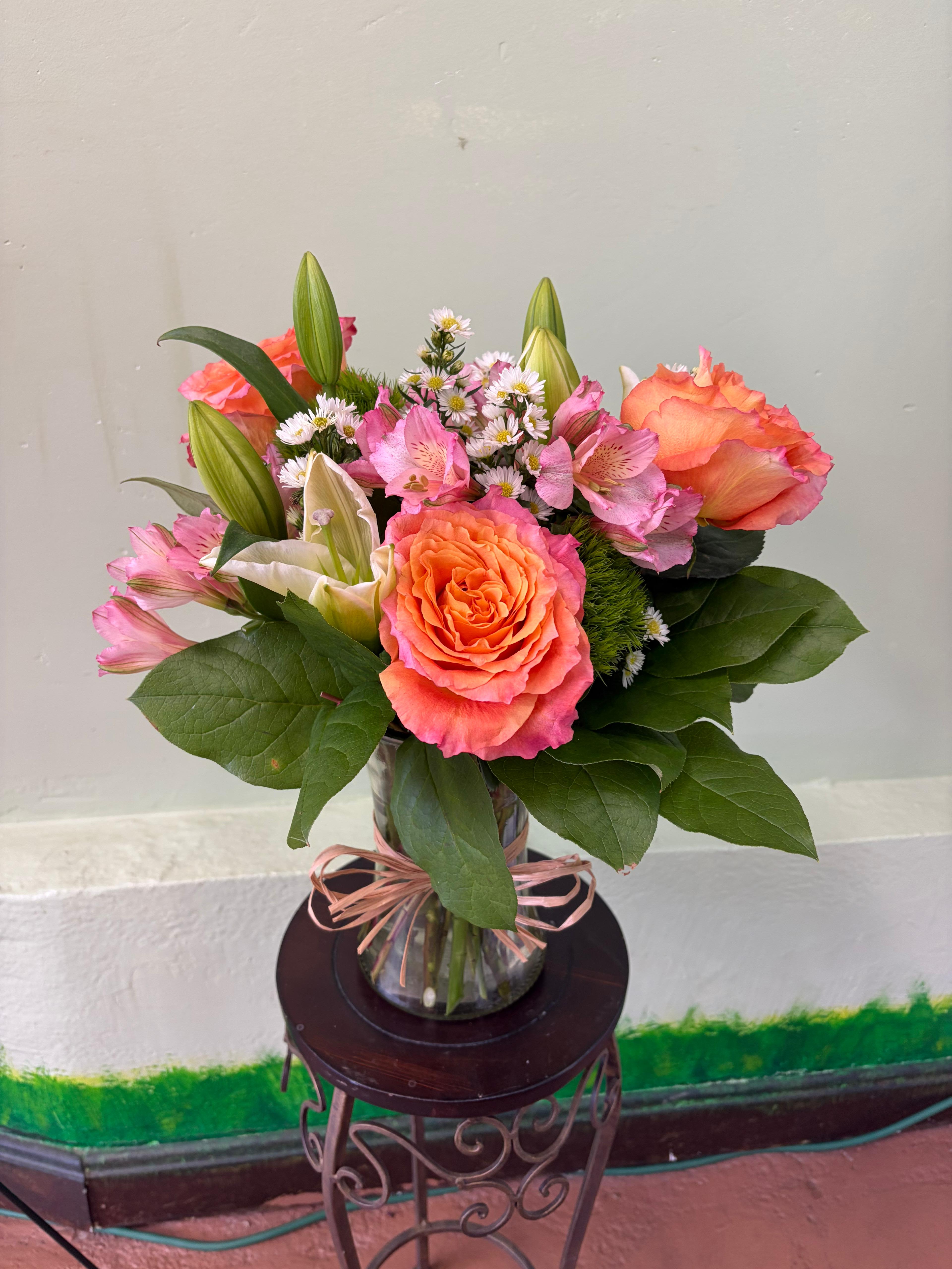 Sunset Reverie  - Capture the fleeting beauty of a twilight sky with our Sunset Reverie. This warm and inviting arrangement features a stunning blend of coral-hued "Free Spirit" roses and delicate pink alstroemeria, designed to bring a touch of a sun-kissed garden into your home. Accented with the soft elegance of white lilies and whimsical feverfew, this arrangement is finished with a rustic raffia bow for a charming, hand-picked feel. It's the perfect choice for expressing gratitude, celebrating a birthday, or simply brightening a loved one's day. 