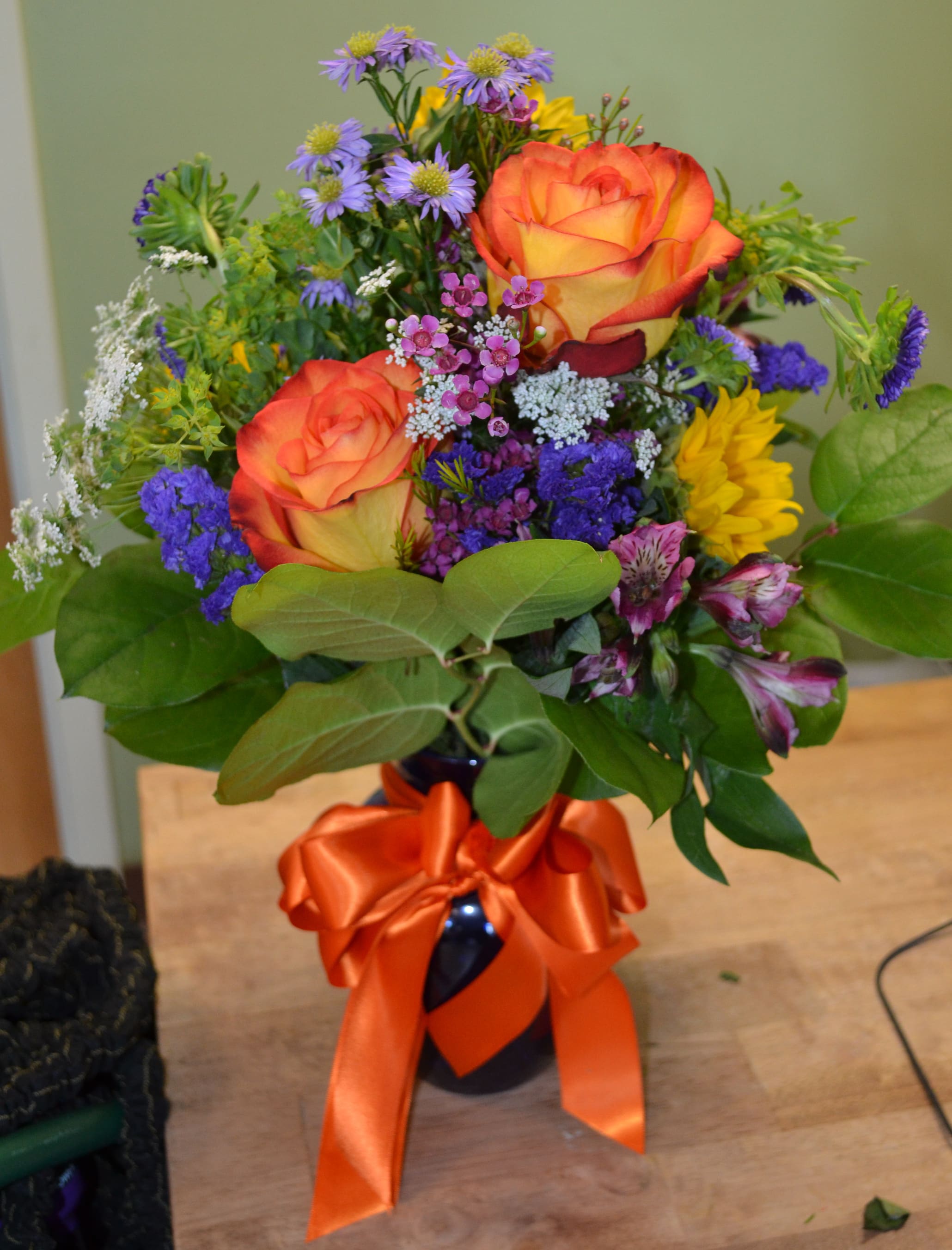 Bouquet of orange roses, purple flowers, and yellow blooms in a dark vase with an orange ribbon