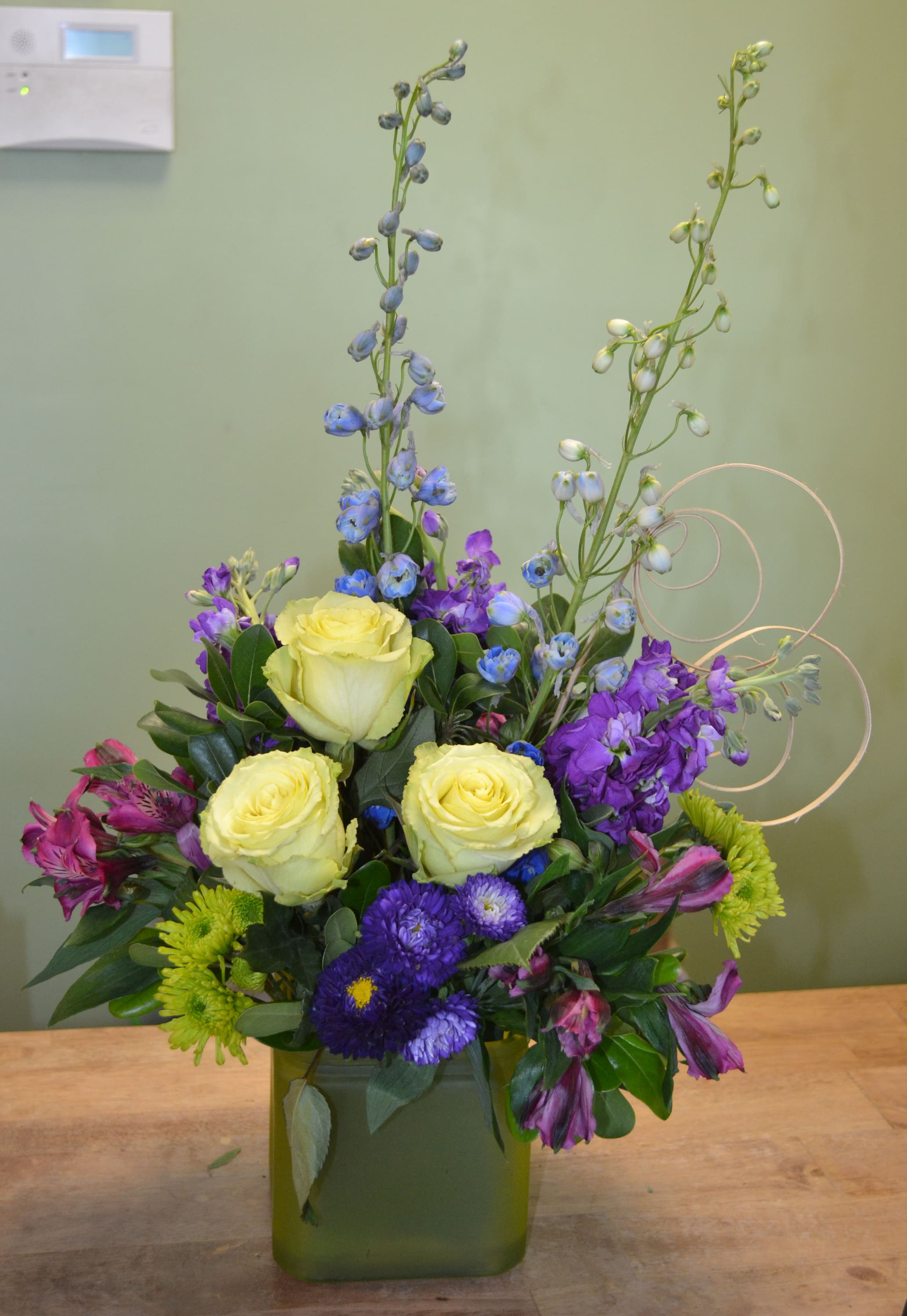 Mixed bouquet with yellow roses, purple flowers, and blue delphinium in a green vase