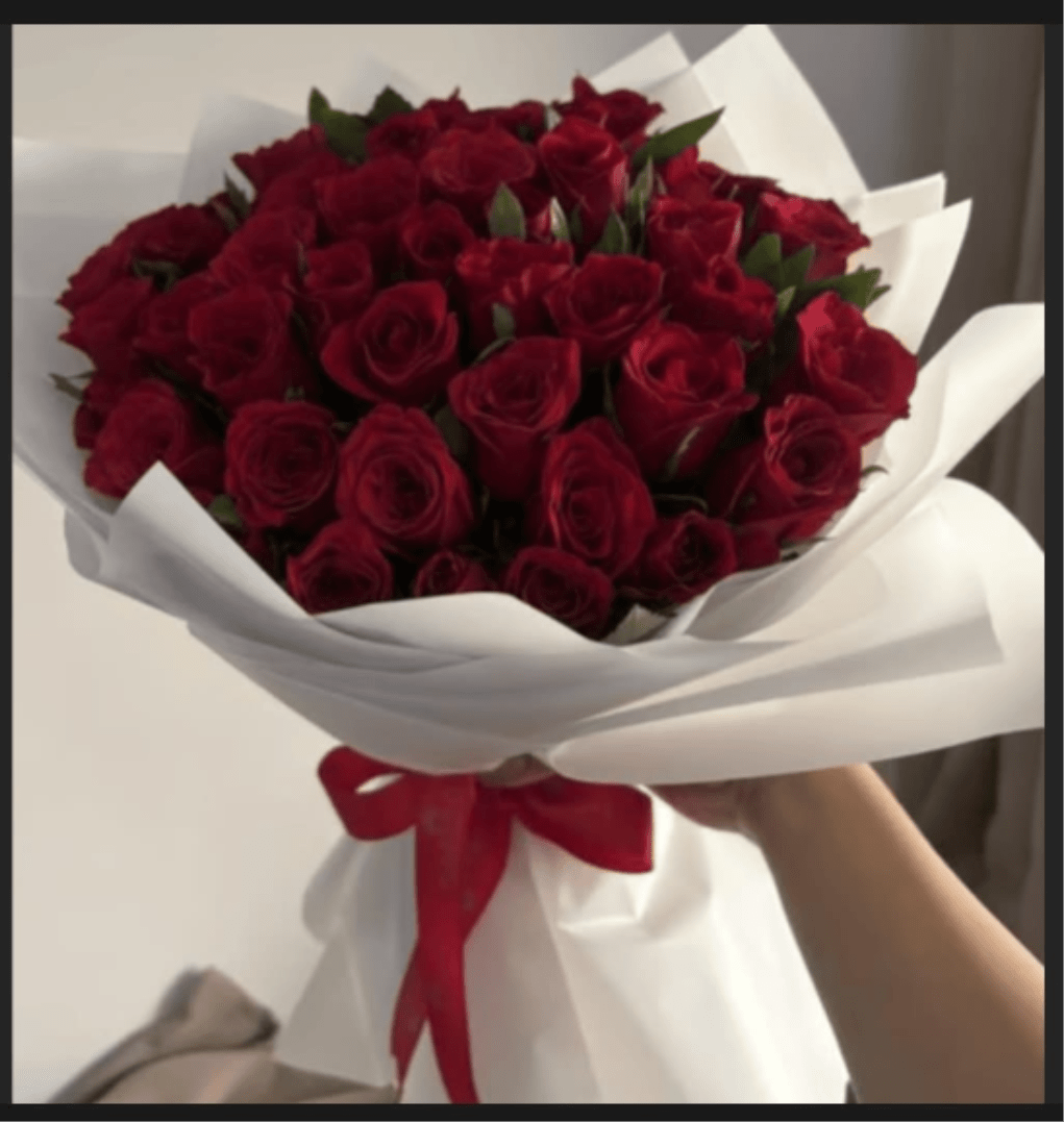RED ROSES BOUQUET - A lush, rounded cluster of deep red roses makes an immediate romantic statement - a red roses bouquet perfect for anniversaries, Valentine's Day, or any moment you want the person you're thinking of to feel truly cherished. The rich crimson petals read as timeless and passionate, while the crisp white paper wrap and glossy red ribbon give the arrangement a polished, gift-ready finish that arrives feeling intentional and elegant.  This medium-to-large hand-held bouquet is densely gathered for a full, abundant look that reads luxurious from every angle. Green foliage peeks subtly between the blooms to lift the color and add fresh contrast. Wrapped in white paper and tied with a red satin ribbon, the presentation keeps the focus on the velvety roses and their striking red-and-white contrast.  Send this bouquet to celebrate a milestone, say "I love you," or make an ordinary day feel special - she'll feel seen and adored when handed this classic arrangement. Ordered for hand delivery, it arrives ready to present; keep the stems in water and trim them slightly on arrival to extend vase life.