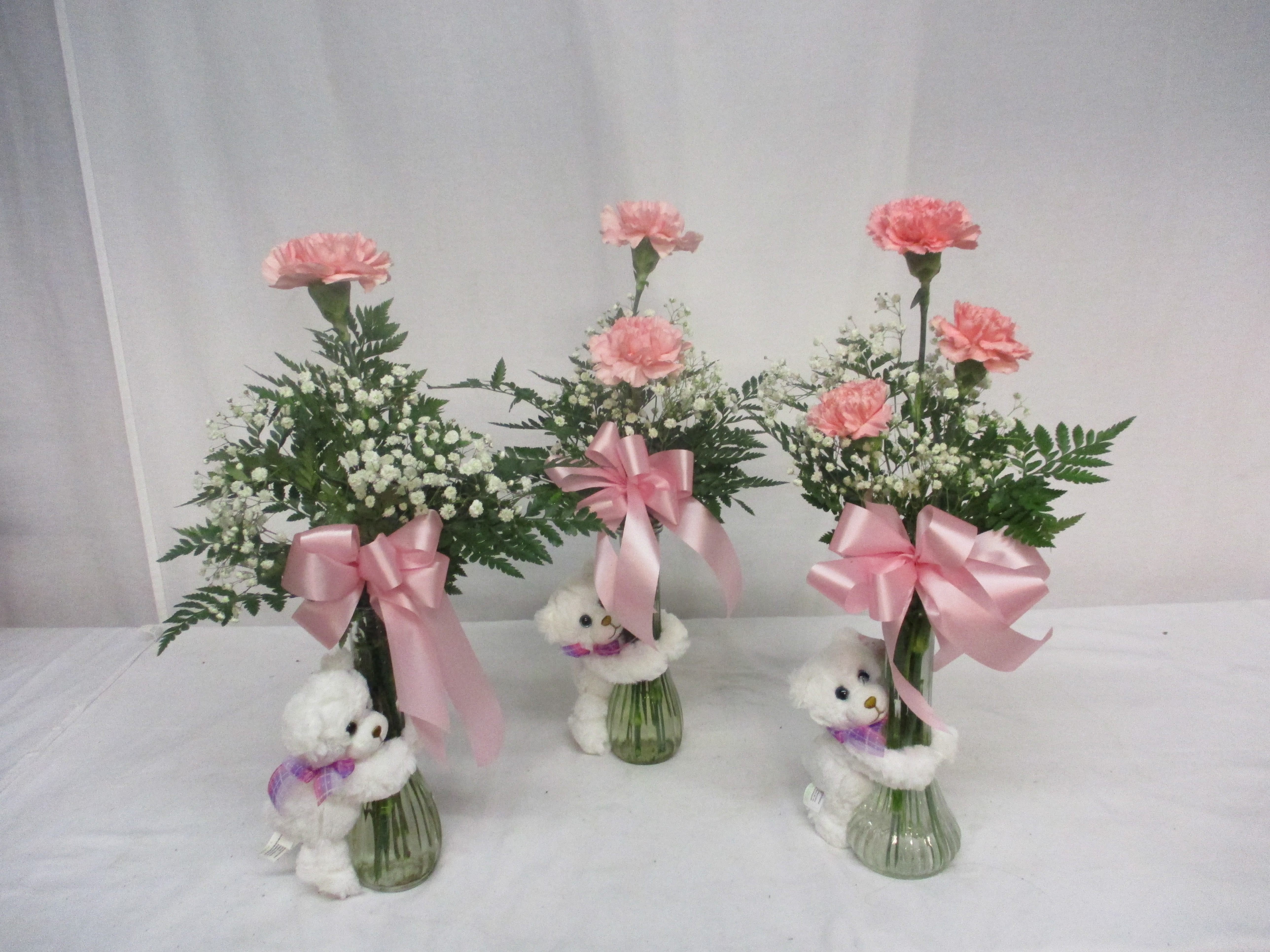 Three pink carnation arrangements in glass vases with teddy bears and ribbons