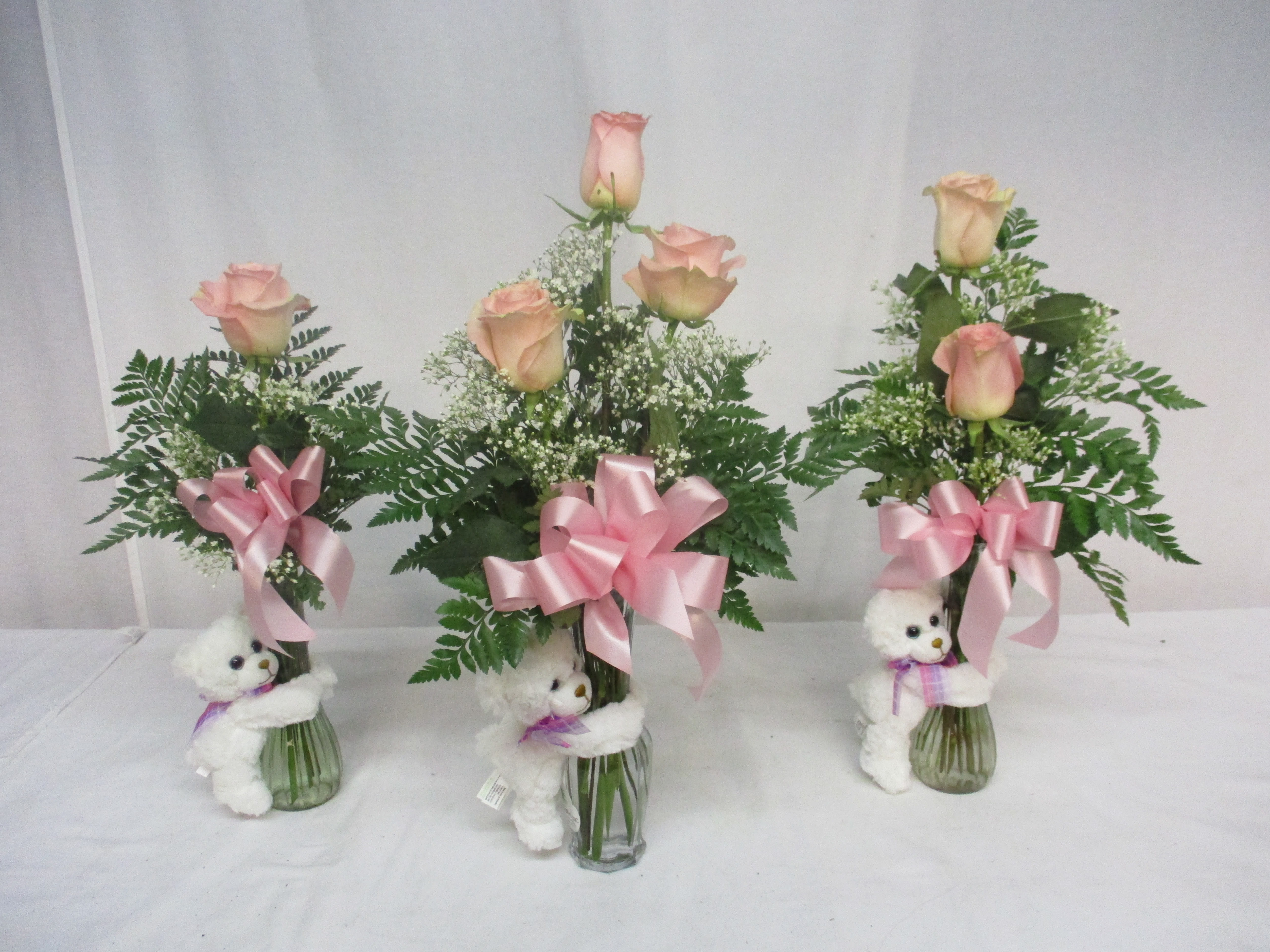 Three pink rose arrangements in glass vases with teddy bears and ribbons