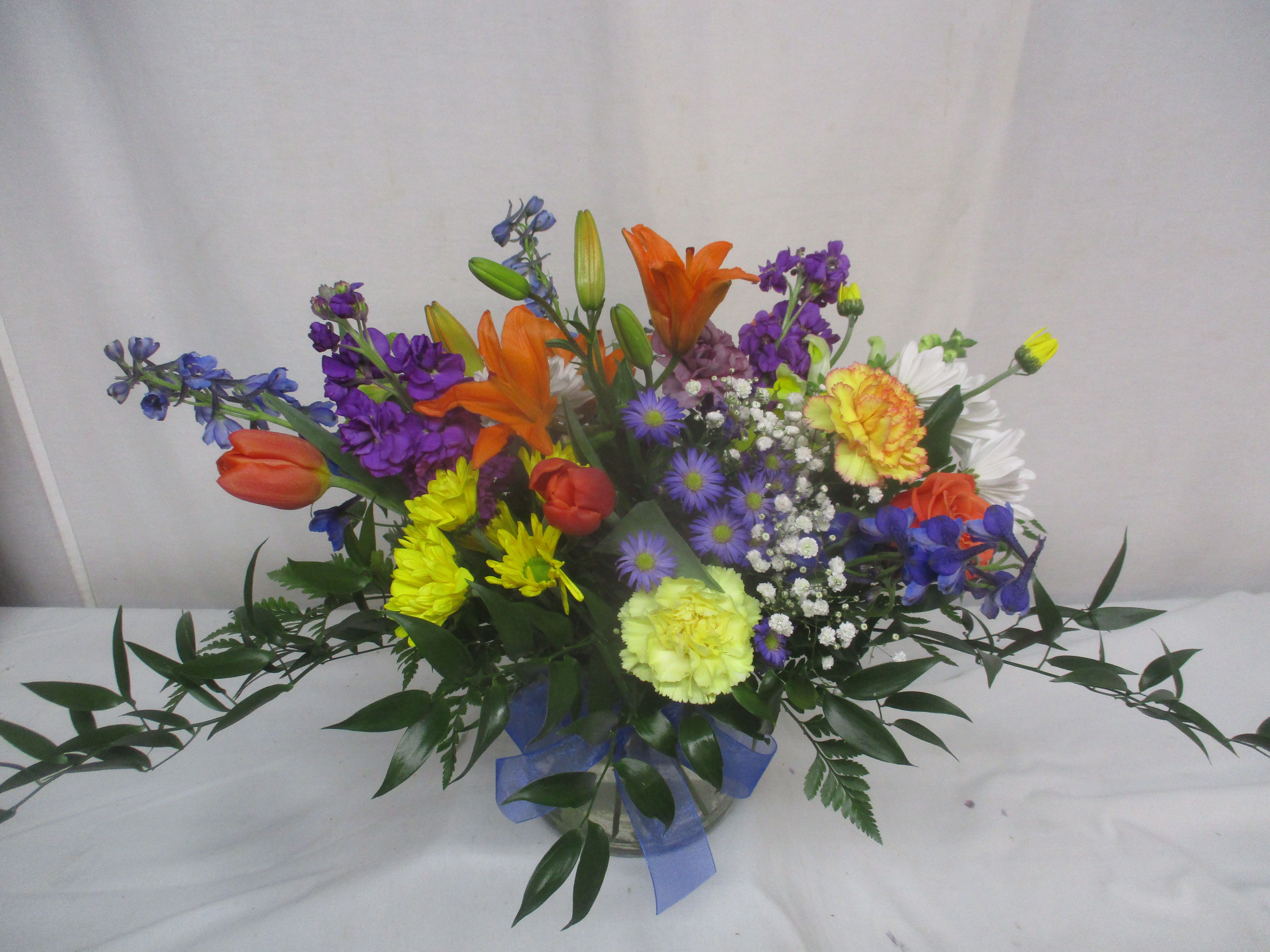 Fields in Bloom - A lively cascade of orange tulips and lilies greets the eye first in this mixed color flower arrangement-perfect as a birthday bouquet, a bright thank-you gift, or an everyday celebration. Fields in Bloom carries that garden-fresh energy into any room with cheerful contrasts of warm orange against cool purples and sunny yellows, so the person you're thinking of will feel genuinely celebrated the moment it arrives.  This wide, low arrangement sits in a clear glass vase tied with a simple blue ribbon, arranged densely with tulips, lilies, daisies, carnations, alstroemeria, asters, stock and delicate baby's breath for soft texture. Long trailing greenery and outward-reaching stems give the design an airy, spread-out silhouette that reads like a freshly gathered patch of spring flowers. Colors include vibrant orange, purple, yellow, white and touches of blue for a bright, rainbow effect.  Ideal for birthdays, congratulations, or sending cheerful get-well wishes, Fields in Bloom is designed to look full and garden-like on a table or mantel without towering over the space. The low vase makes it easy to place anywhere; refresh water every other day and trim stems if needed to keep blooms lively for as long as possible.