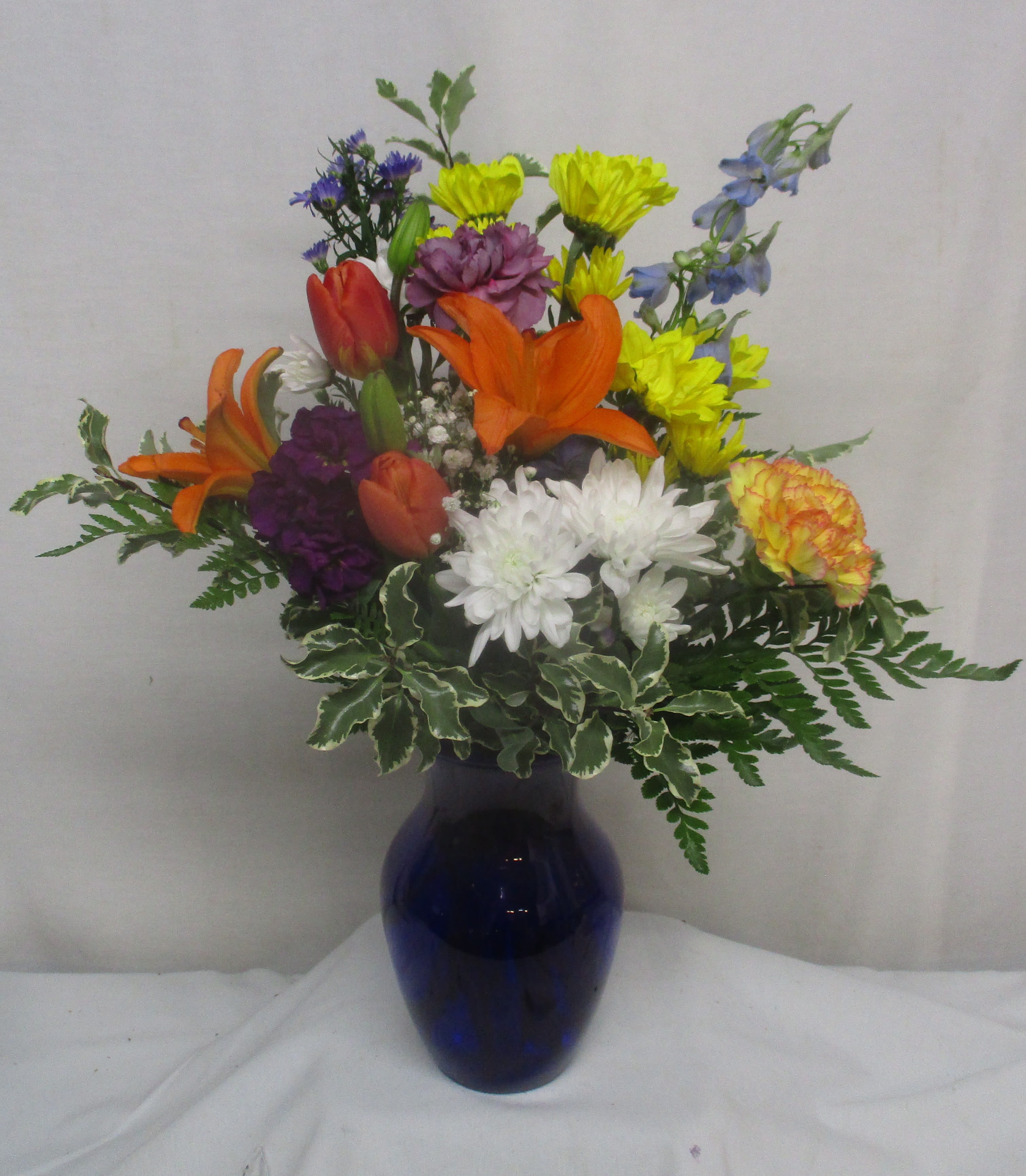 Happy Days Bouquet - Vivid orange Asiatic lilies and fiery red-orange tulips take center stage in this mixed-color flower arrangement, making the Happy Day Bouquet an ideal birthday bouquet or congratulations gift. The lively combination of yellow daisy-like blooms and crisp white chrysanthemums adds sunny contrast while purple accent flowers lend a playful pop - the kind of bright, joyful display that lets the person you're thinking of know you celebrate them.  Arranged in a cool blue glass vase, the rounded, upright design feels full and classic without being fussy. Textures range from the smooth petals of the tulips and lilies to the soft, pompon-like chrysanthemums, creating visual interest from every angle. This everyday bouquet suits thank-you gestures, cheerful get-wells, or simply to make someone feel genuinely seen; sizing is a full, table-friendly arrangement that arrives ready to display in the included blue vase.