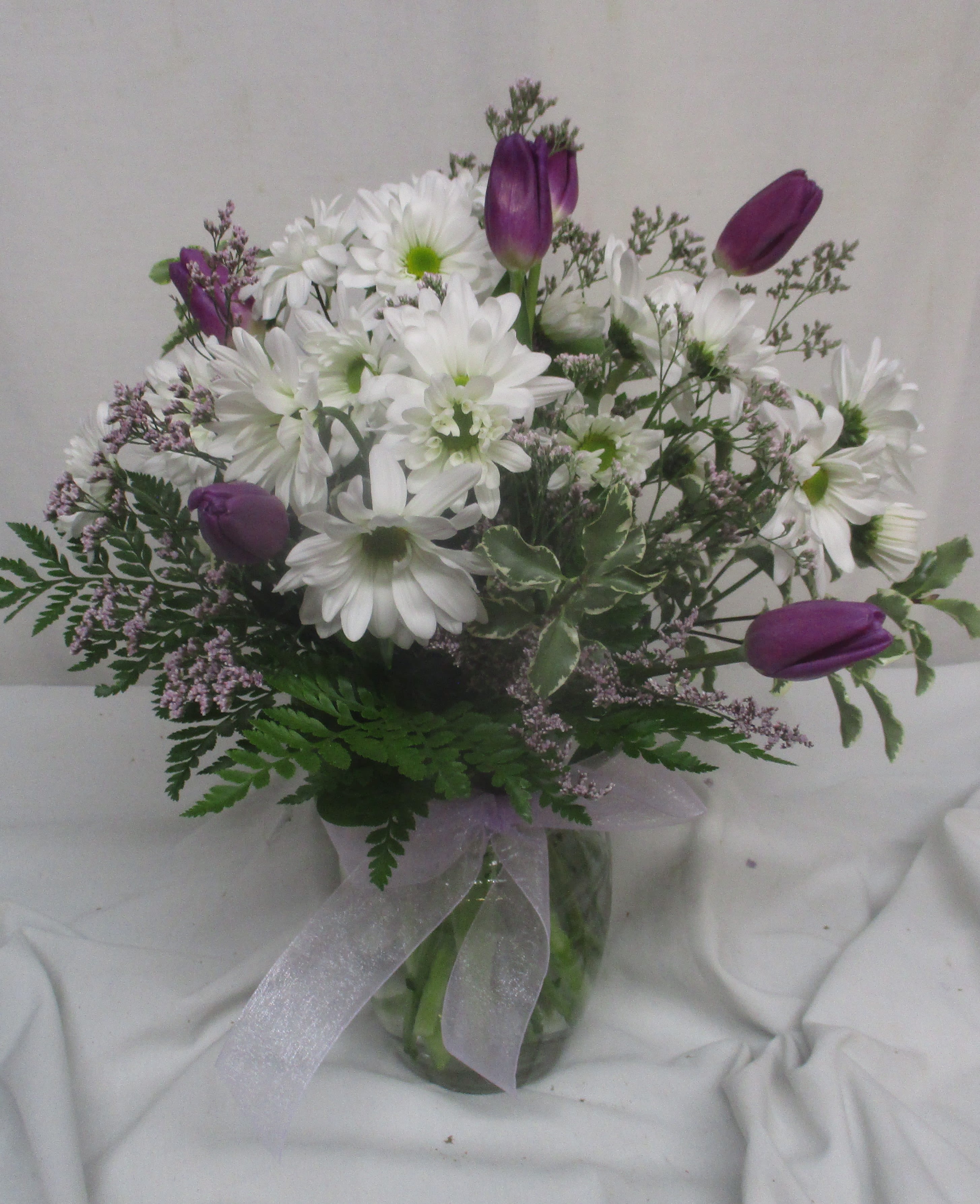 Tulip Time - Purple tulips and white daisy-like blooms create a fresh, garden-style statement-perfect as a spring bouquet or a cheerful everyday arrangement. This flower arrangement is a great choice for birthdays, Mother's Day, or to simply let the person you're thinking of know they're appreciated; the soft colors and airy silhouette feel both joyful and gentle on the recipient.  Gathered in a clear glass vase and finished with a pale sheer ribbon, the rounded, loose design mixes purple tulips, white daisies, fern-like foliage, and airy filler for natural texture and movement. The compact, hand-tied shape sits nicely on a table or mantel and arrives ready to display-no extra work required-so it's an effortless, thoughtful gift that will make someone feel genuinely seen.