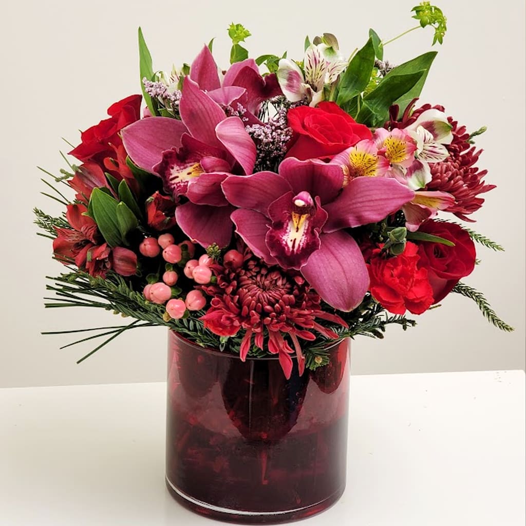 Cozy Comfort - The Cozy Comfort bouquet comprises an array of vivid red blooms, luscious greens, and striking beauty to make your loved one’s day. With rich burgundy cymbidium orchids, vibrant red blooms, and delicate pink accents.   Presented in a matching dark red vase, this bouquet makes a striking statement, perfect for conveying deep passion, congratulations, or as a centerpiece that captures the essence of opulent beauty.  Approximately 16"H x16" W. 