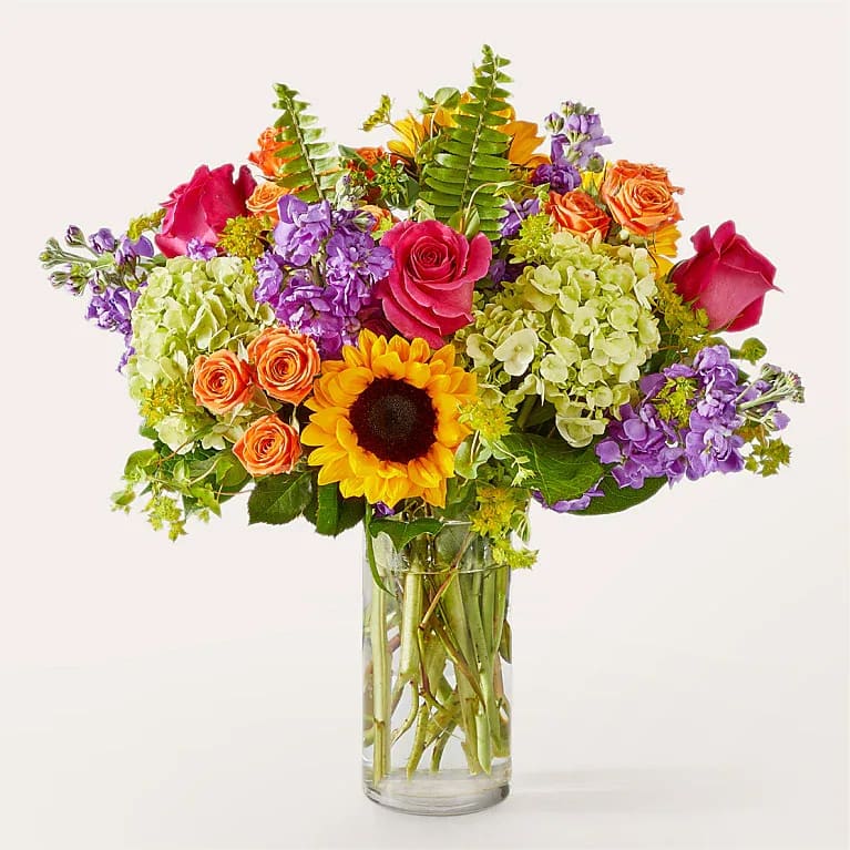 Colorful bouquet of roses, sunflowers, hydrangeas, and purple blooms in a glass vase