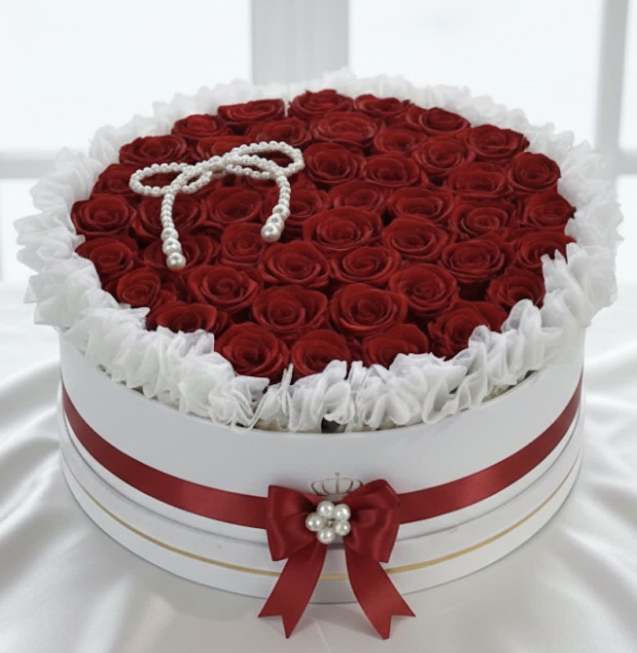 Round box filled with red roses and white ruffled trim, topped with a pearl bow