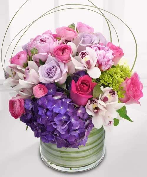 Low round arrangement of pink and lavender blooms with orchids and purple hydrangea in a glass vase