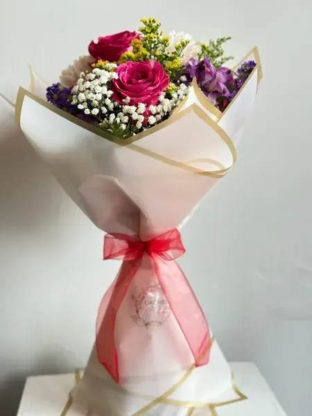 Bouquet of pink roses, purple flowers, and white filler wrapped in paper with a red ribbon