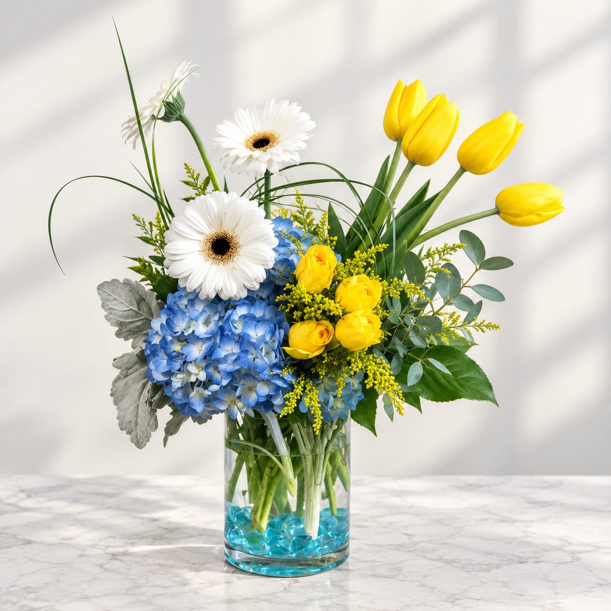 Bouquet of yellow tulips, white gerbera daisies, blue hydrangea, and yellow roses in a glass vase