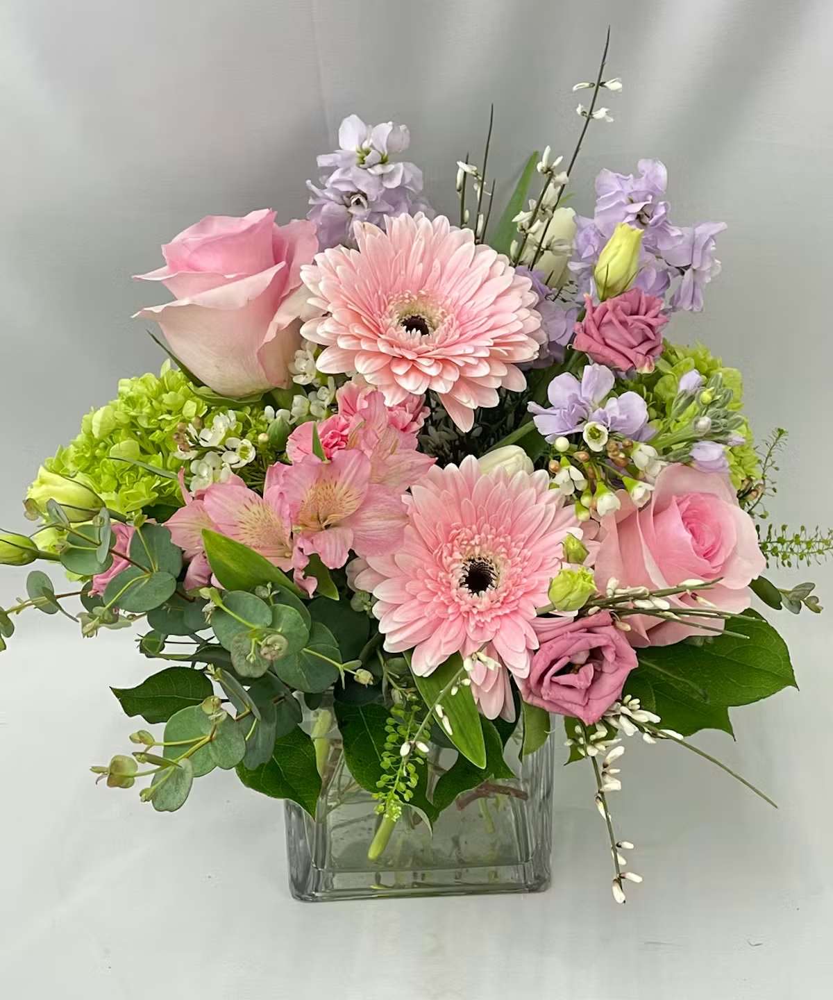 Blushing Meadow - Soft pink roses, delicate gerbera daisies, lavender blooms, and fresh green hydrangea come together in this charming garden-style arrangement. Designed in a modern glass cube vase with lush greenery and airy accents, it has a light, cheerful feel that is perfect for celebrating a birthday, welcoming spring, or sending a thoughtful reminder that someone is loved.  Substitutions may be made depending on availability of flowers.