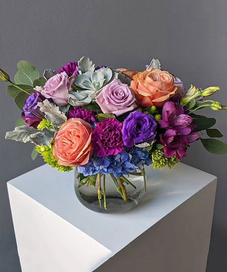 Twilight Garden - A lush and artistic arrangement featuring lavender and peach roses, vibrant purple blooms, blue hydrangea, and accents of fresh greenery and succulents. Designed in a modern glass vase, this striking piece blends rich jewel tones with soft pastel shades for a look that feels both elegant and whimsical. Perfect for birthdays, anniversaries, celebrations, or adding a beautiful statement to any room.  Please note: Due to seasonal and supplier availability, certain flowers or accents may be substituted with blooms of equal or greater value while preserving the overall style, color palette, and fullness of the arrangement.
