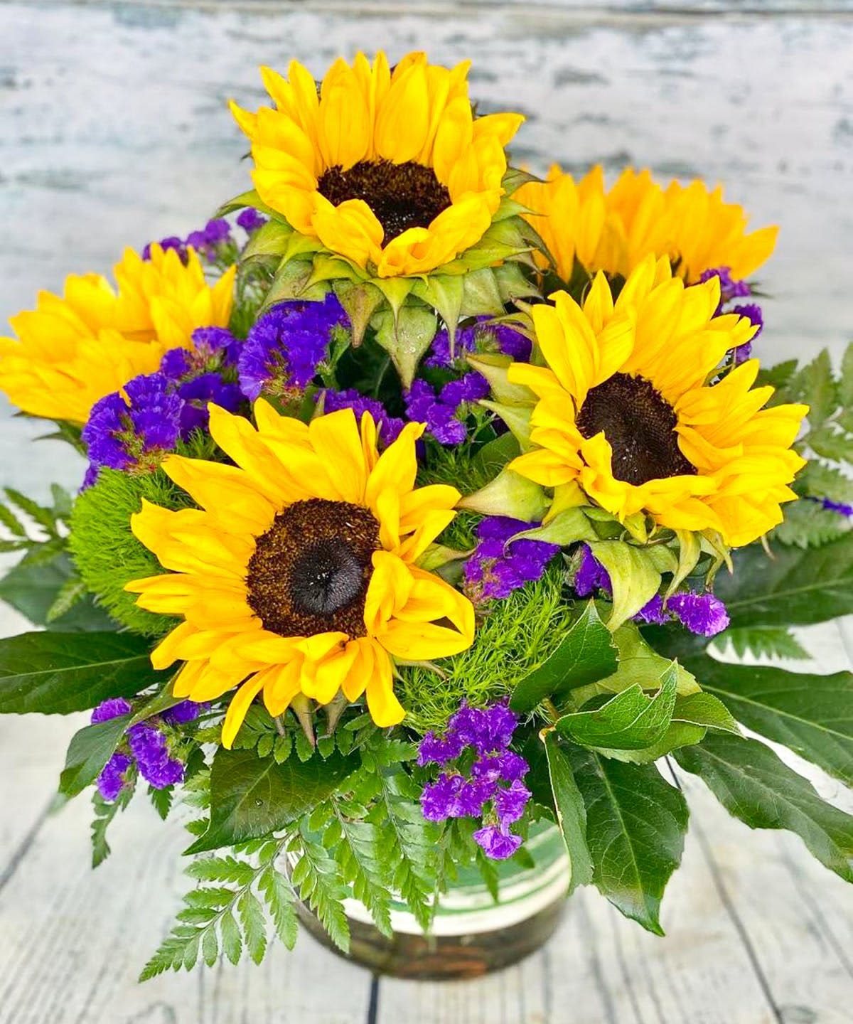 Sunflower Serenade - Bright and cheerful, Sunflower Serenade features radiant sunflowers accented with vivid purple statice and fresh seasonal greenery in a clear glass vase. This bold and sunny arrangement is perfect for birthdays, get well wishes, thank you gifts, or simply bringing a little happiness to someone’s day.  Please note: Flower varieties and accent flowers may vary based on seasonal availability. Substitutions may be made with flowers of equal or greater value while preserving the overall style, color palette, and feel of the arrangement.