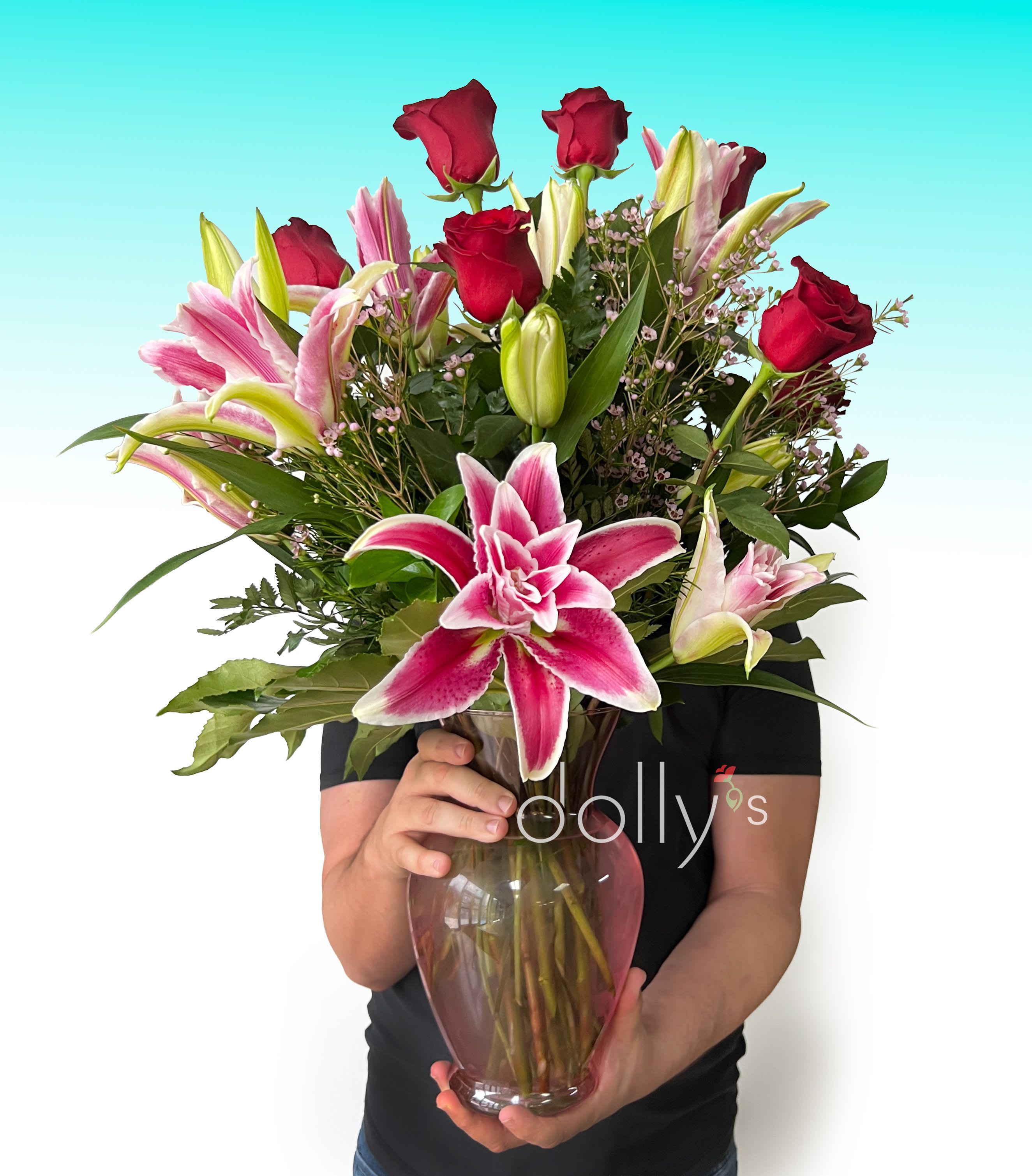 Ruby Muse - Deep red roses rising in a tall, elegant cluster meet large pink lilies-some in full bloom and others coyly in bud-to create a romantic, vertical bouquet perfect for occasions like anniversaries, Valentine's Day, Mother's Day, or a meaningful birthday. The combination of red, pink, and touches of white reads as both passionate and tender, the kind of arrangement that tells someone you love them, are sorry, or are gratefully thinking of them.  Set in a tall glass vase that subtly echoes the pink tones of the blooms, this elongated design emphasizes height and graceful lines. Dense foliage and small filler blossoms wrap around the roses and lilies for rich texture and a lush, full look; the varying lily stages add movement and scent to the composition. Ideal for expressing love and romance, saying "I'm sorry," offering thanks, or simply brightening someone's day-especially around Mother's Day or Breast Cancer Awareness Month-this arrangement arrives ready to display and makes a striking focal point in an entryway or dining room.