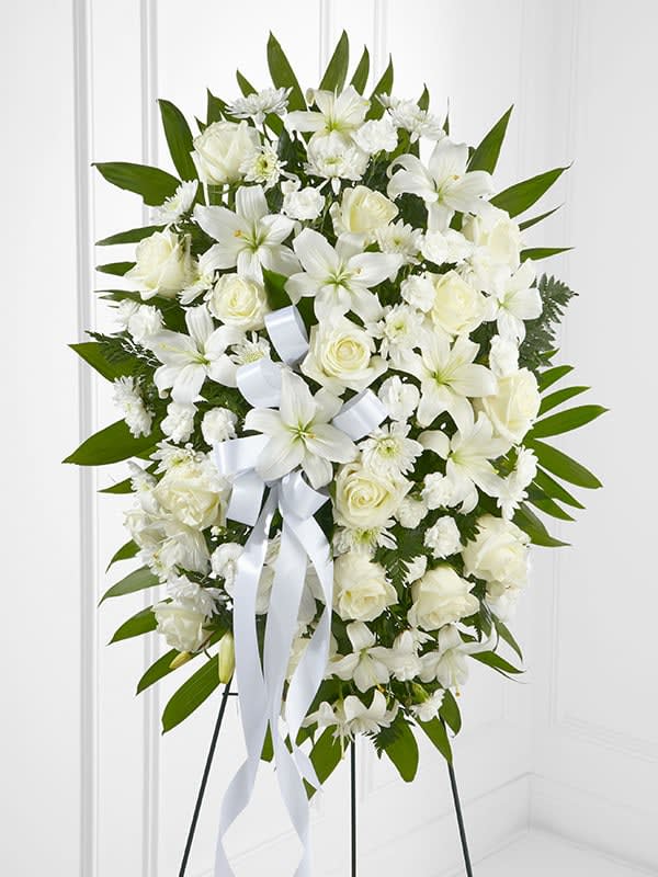 Standing spray of white lilies, roses, and carnations with a white ribbon on an easel.