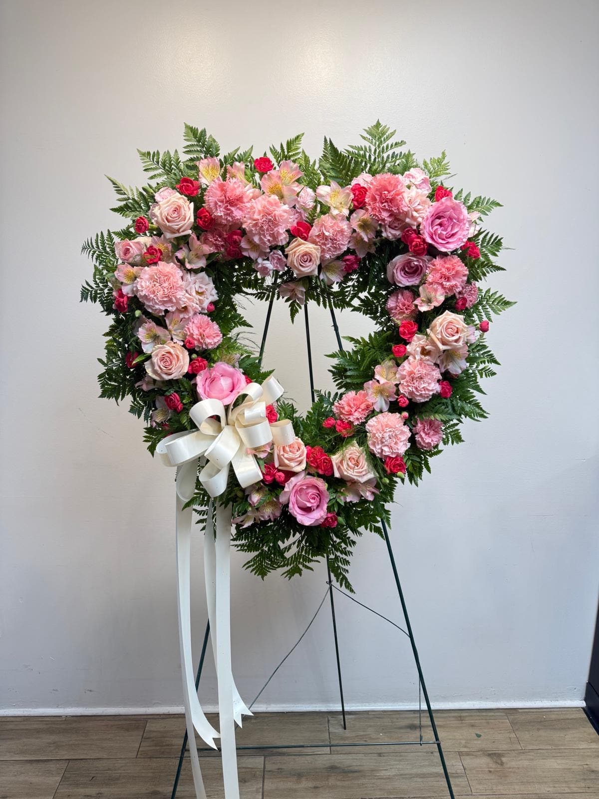 Heart-shaped pink floral wreath on a stand with a white ribbon bow