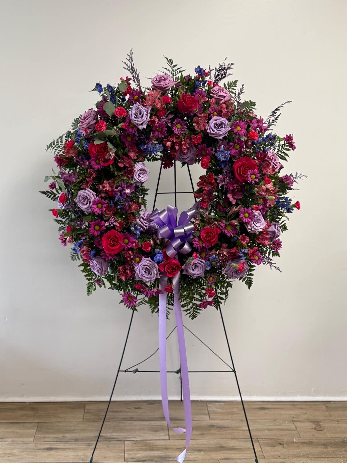Large floral wreath on a stand with pink, purple, and blue flowers and a lavender ribbon
