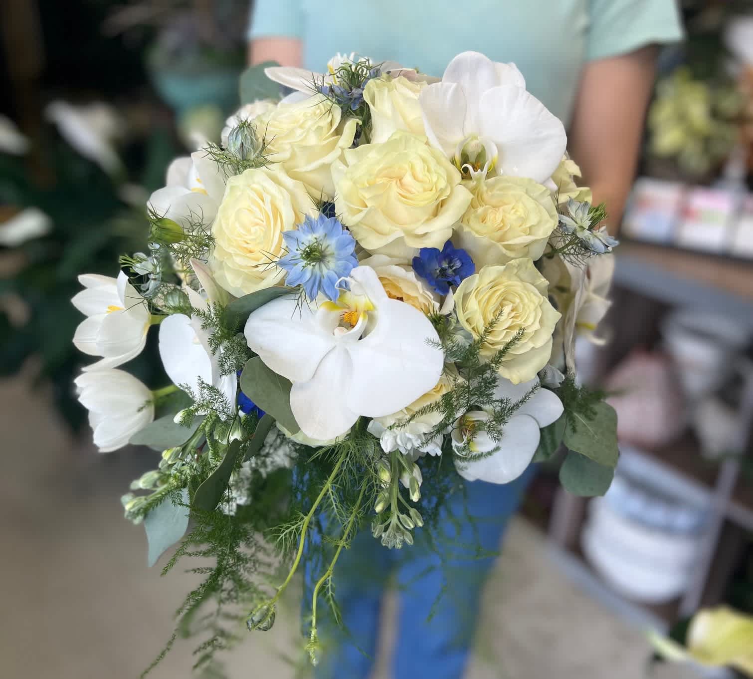 Hand-tied bouquet of cream roses, white orchids, and blue accent flowers with trailing greenery