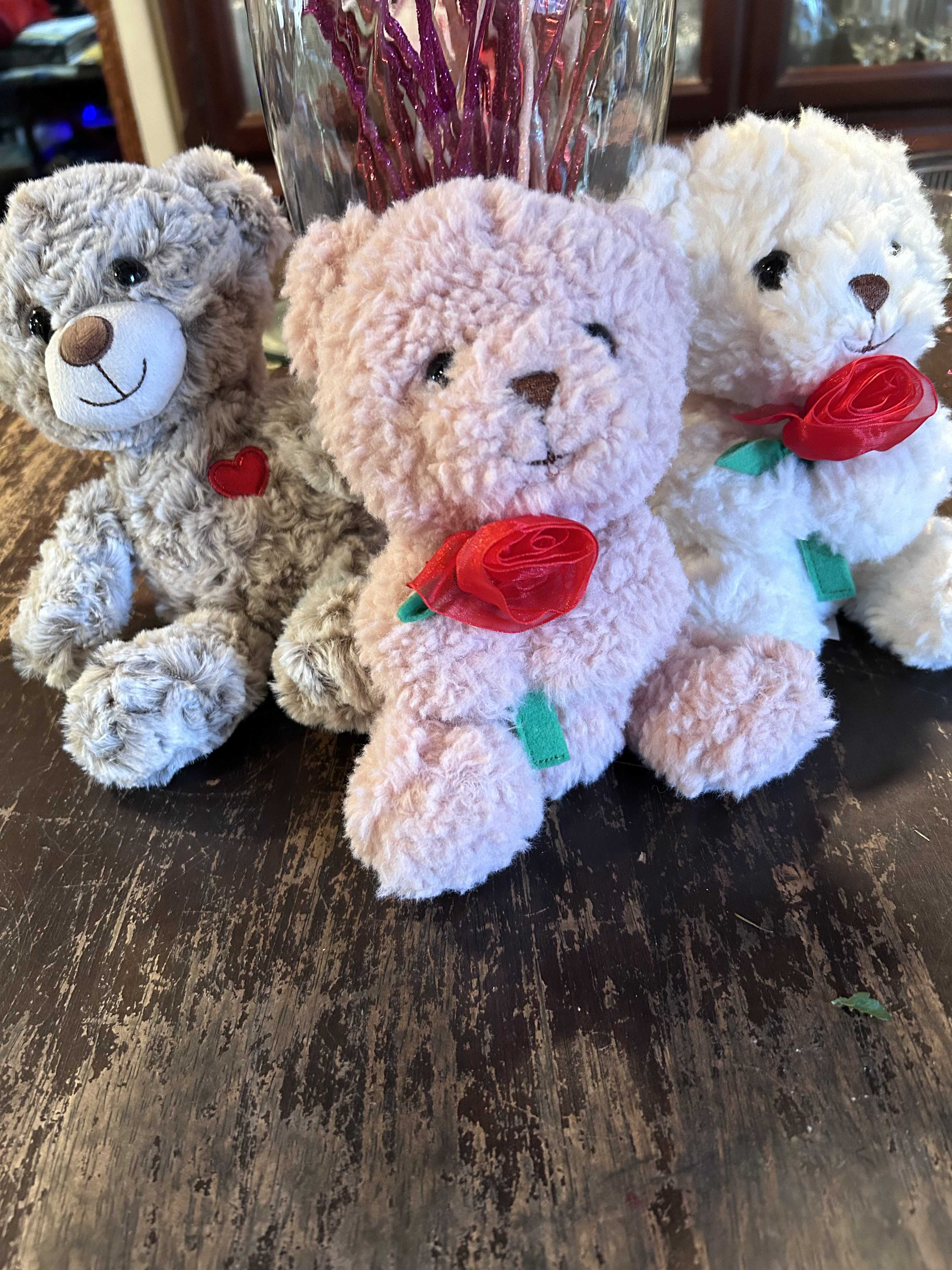 Three plush teddy bears with ribbon roses sit on a table.