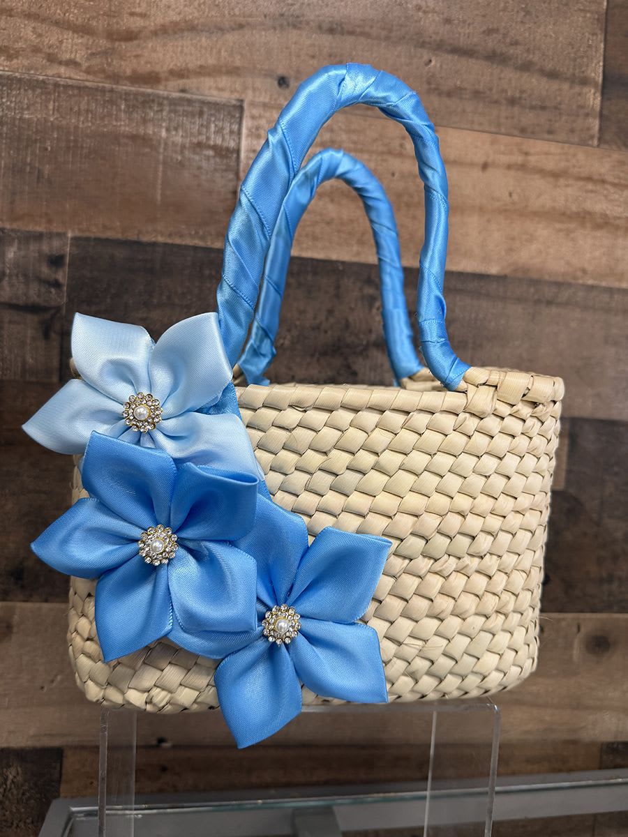 Artisan Purse - Blue Theme - A handcrafted woven straw handbag trimmed with satin ribbon flowers and sparkling rhinestone centers makes a charming gift-ready accessory for a spring baby shower, birthday, or hostess present. This small woven basket-style arrangement reads like a polished keepsake rather than a traditional bouquet-its bright blue and pale blue satin blooms (with jeweled centers) or the softer ivory, champagne and gold ribbon flowers bring instant cheer and a touch of couture to the recipient's moment.  Each handbag features natural straw texture, two wrapped handles, and decorative satin ribbon flowers finished with rhinestone brooch centers for a playful yet elegant look. Sizes are compact and structured-perfect for pairing with small gifts, note cards, or a single posy if the recipient prefers-even when no fresh flowers are included the piece feels celebratory. Choose the colorway that suits the occasion: bold red flowers for a festive pop, bright blue for a spring or baby-themed surprise, or neutral champagne and ivory for a more formal gift.
