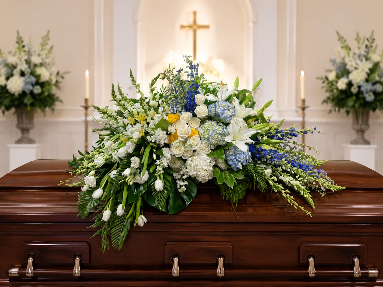 Watercolors Casket Piece by Petal Perfection  - Soft blue delphinium and round blue hydrangeas create a serene horizontal display that brings calm to difficult moments - a sympathy and funeral arrangement designed to comfort the person you're remembering. This low casket-style flower arrangement pairs clusters of white roses and fragrant white lilies with white tulips and small white blooms, their graceful, cascading edges evoking gentle movement and quiet elegance.  Set in a low container, the Watercolors piece measures as an elongated, table-friendly centerpiece that also suits a funeral display or a home remembrance. The palette of cool blues, crisp whites and subtle yellow accents gives the composition a refined, restorative feel; textures range from the full, cloudlike hydrangeas to the sculpted rose blooms and the tall, airy delphinium. Order knowing this arrangement arrives ready to place - its horizontal profile makes it easy to situate on a casket spray area, dining table, or mantel, and the mix of long-stemmed and clustered flowers holds visual presence without overwhelming the space.