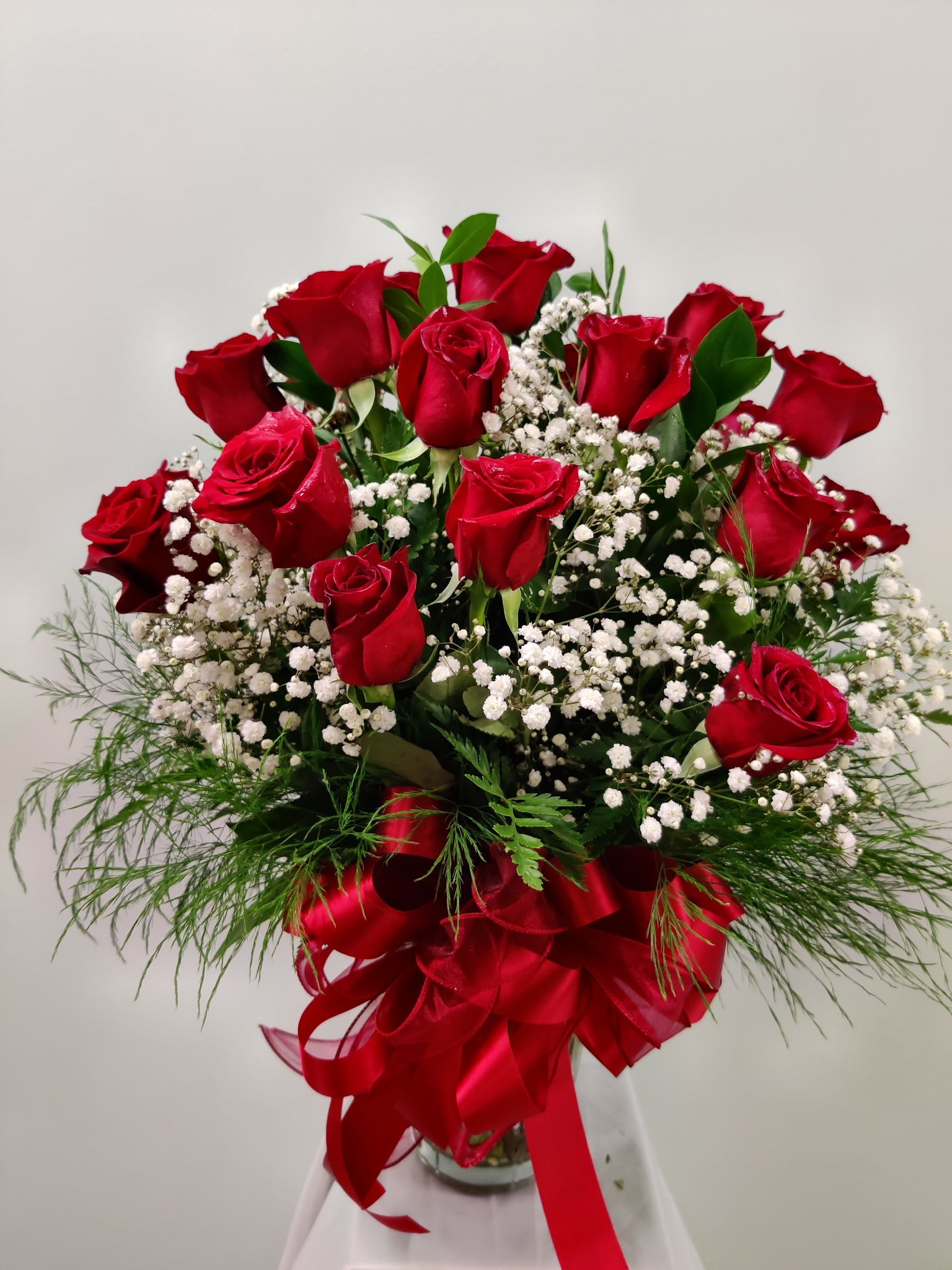 Double the Love - A generous cluster of two dozen red roses rises above a cloud of delicate white filler blooms for a bold, classic statement-perfect as an anniversary bouquet, Valentine's Day surprise, or a "love and romance" gesture that makes someone feel deeply seen. This red rose arrangement also reads respectfully for sympathy, Memorial Day, or a heartfelt "I'm sorry," while equally fitting celebratory moments like birthdays, graduations, or Mother's Day.  Arranged in a clear glass vase so the long stems and fresh greenery show through, the design balances rich crimson petals with crisp white accents for striking contrast. A large, multi-looped red satin ribbon bow at the base adds formal polish and romantic flair. Sized as 24 long-stem roses, this arrangement delivers strong visual impact and lasting presence on tabletops, mantels, or reception desks.  Send this when you want the recipient to feel honored, comforted, or celebrated-whether it's for Father's Day, a housewarming, thank-you, retirement, or just because. For best results, keep the vase topped up with fresh water and recut stems slightly if you need to refresh the display; delivered ready to enjoy and designed to convey your message with classic elegance.