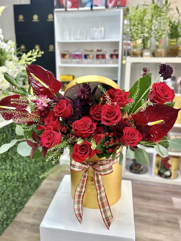 Red roses and dark red tropical blooms arranged in a gold hatbox with a plaid ribbon bow.