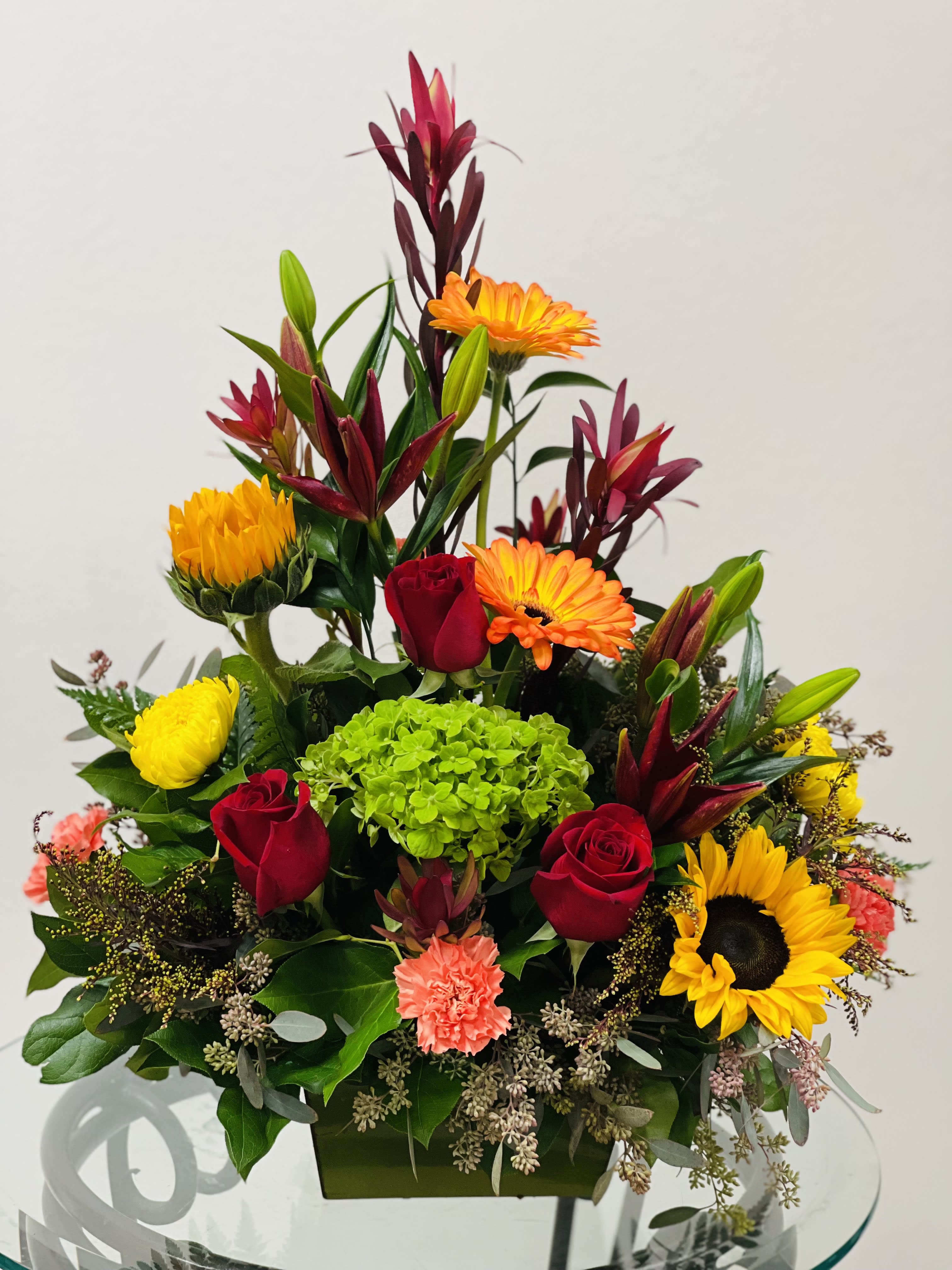 Mixed bouquet with red roses, orange gerberas, yellow blooms, and green hydrangea in a square vase