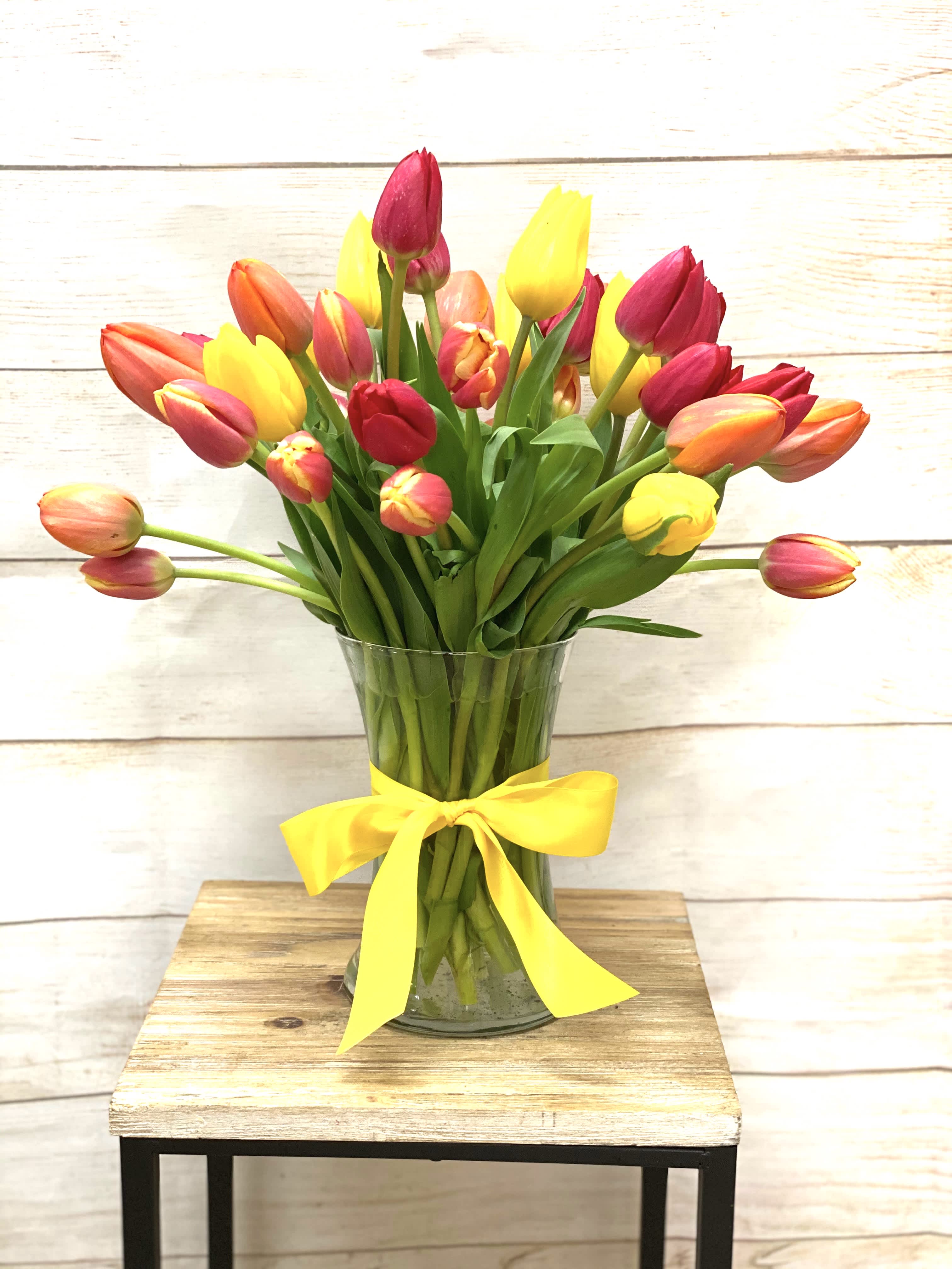 Mixed red, yellow, and orange tulips in a clear glass vase with a yellow ribbon bow.