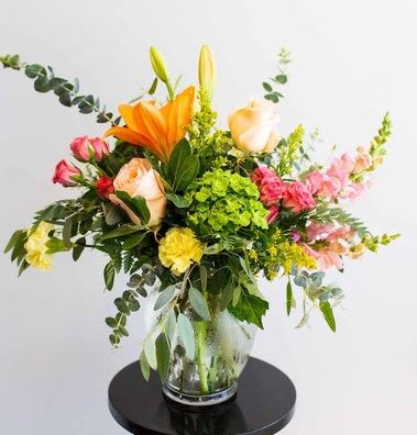 Sun Kissed Blooms - Inspired by the sunrises and sunsets, this asymmetrical arrangement features soft, warm hues of roses, lilies, and carnations. Finished with bright greens and eucalyptus. *** VASE WILL VARY***