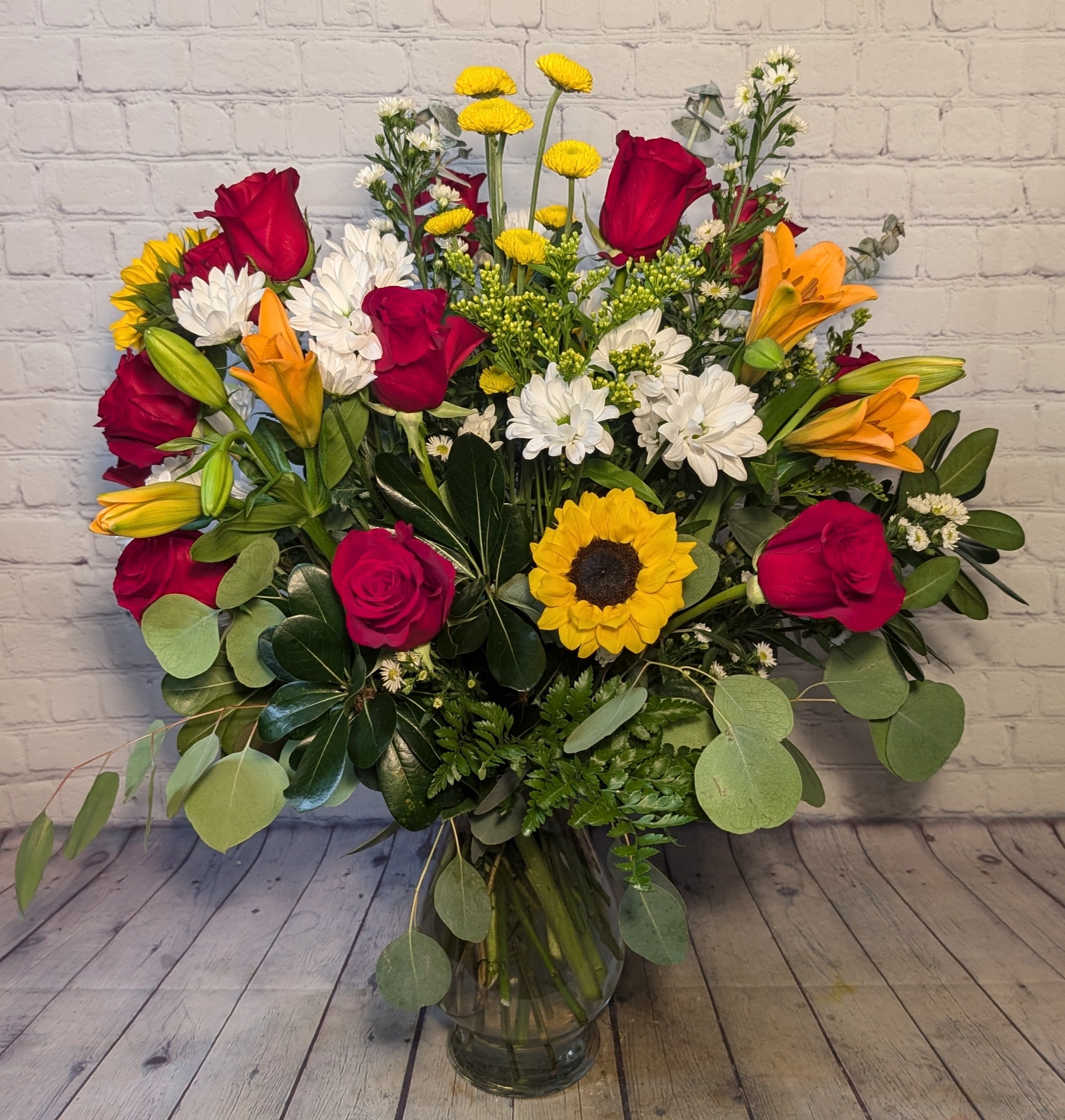 Mixed bouquet of red roses, lilies, white daisies, and a sunflower in a glass vase