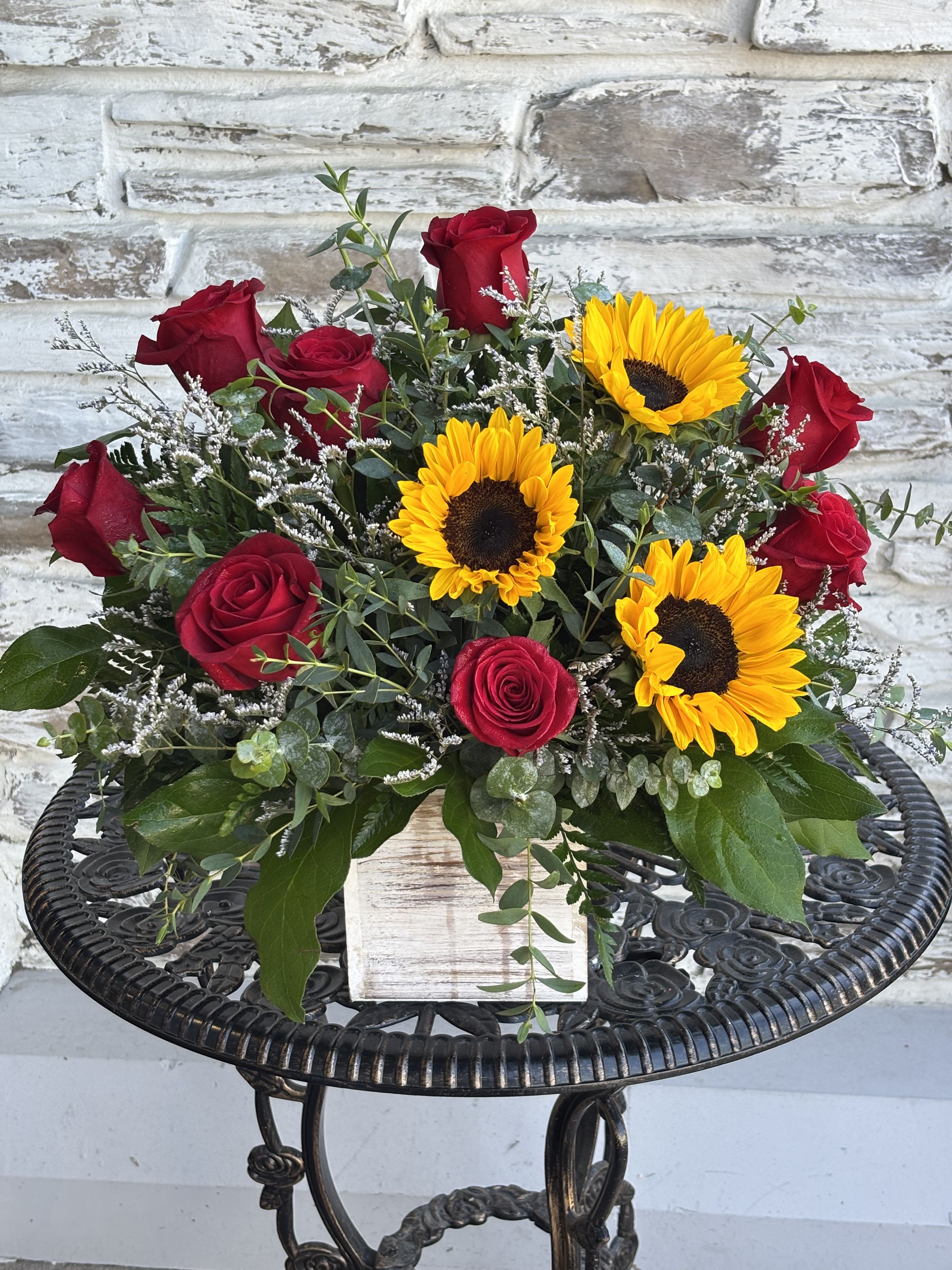 Love In The Sun - This rustic-meets-romantic arrangement blends deep, red roses and vibrant golden sunflowers nestled in a textured wooden box. It tells the story of a love that is both intensely passionate and wonderfully joyful, perfectly balancing fiery romance with unwavering warmth, loyalty, and everyday happiness.