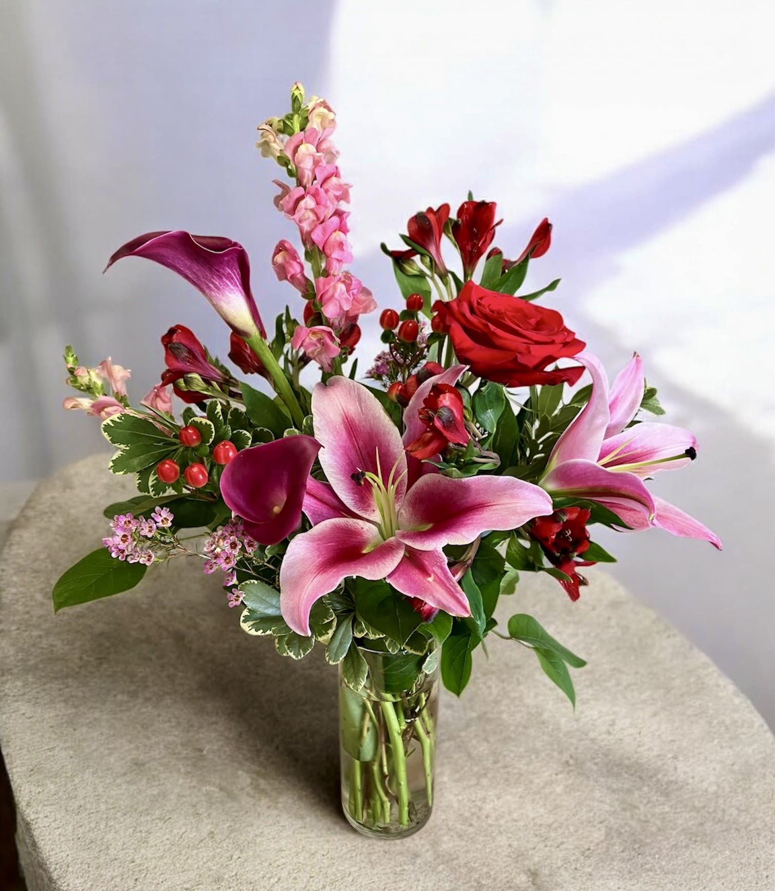 My Beloved - A vibrant and elegant floral arrangement featuring lush pink Oriental lilies as the focal point, accented by rich purple calla lilies and bold red alstroemeria for depth and contrast. Soft pink snapdragons add graceful height and movement, while delicate wax flower brings a light, airy texture. Clusters of red hypericum berries provide a striking pop of color, with variegated pittosporum greenery, this design feels timeless and intimate.
