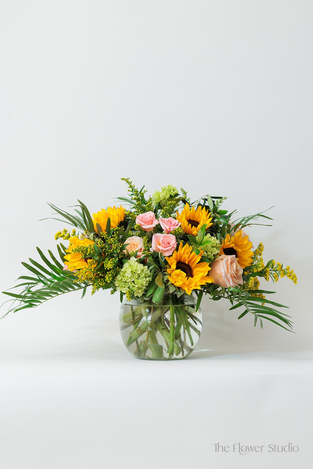 Low round arrangement of yellow sunflowers and pink roses in a clear glass bowl vase