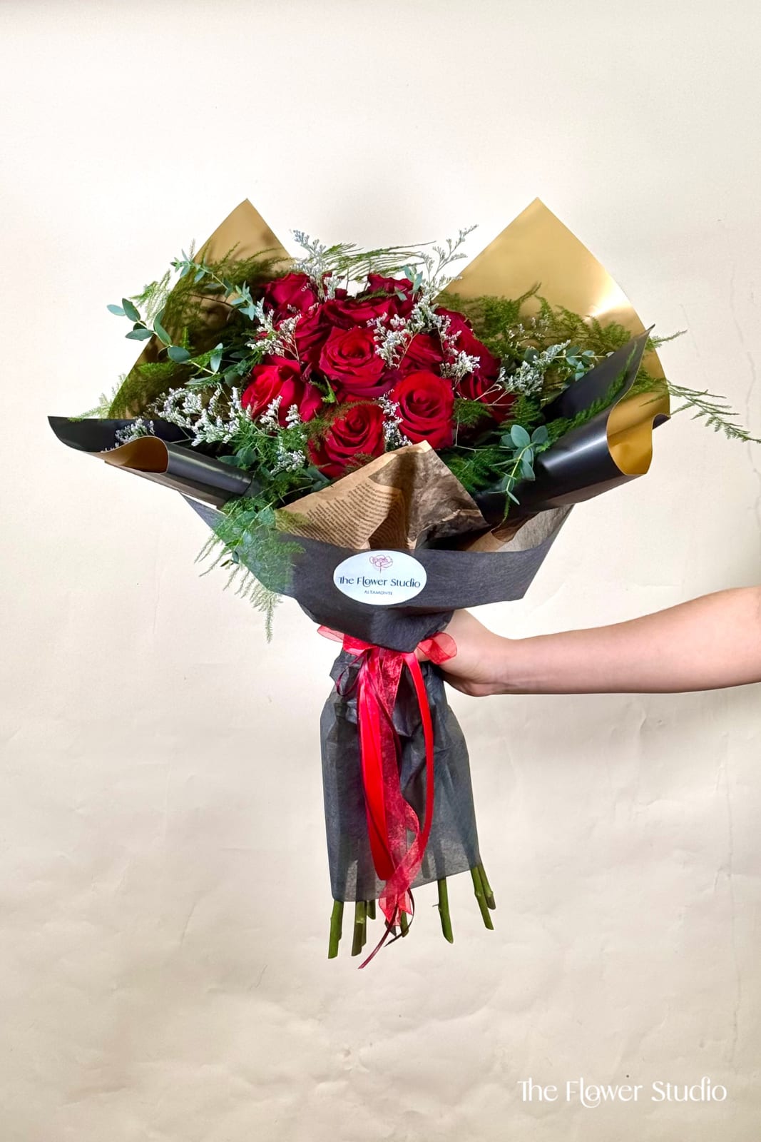 Hand-tied bouquet of red roses with white filler flowers wrapped in black and gold paper and red ribbon
