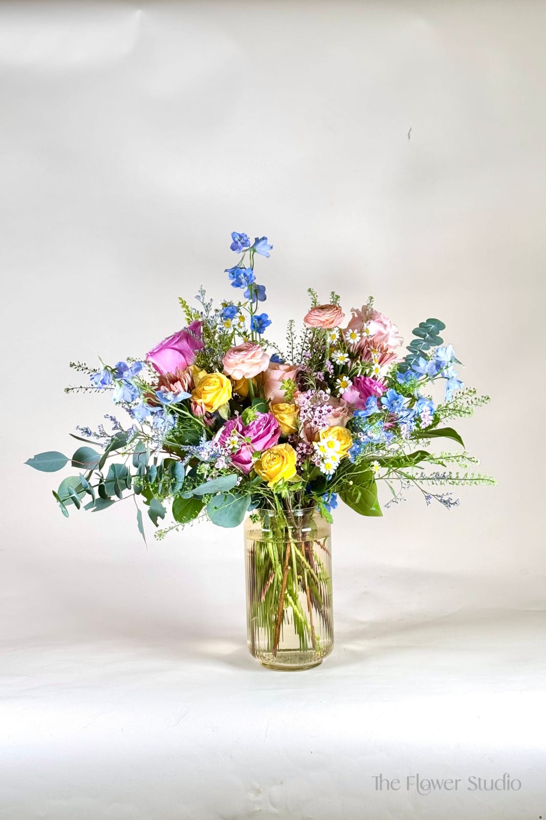 Tall mixed bouquet of pink, yellow, white, and blue flowers arranged in a clear glass vase
