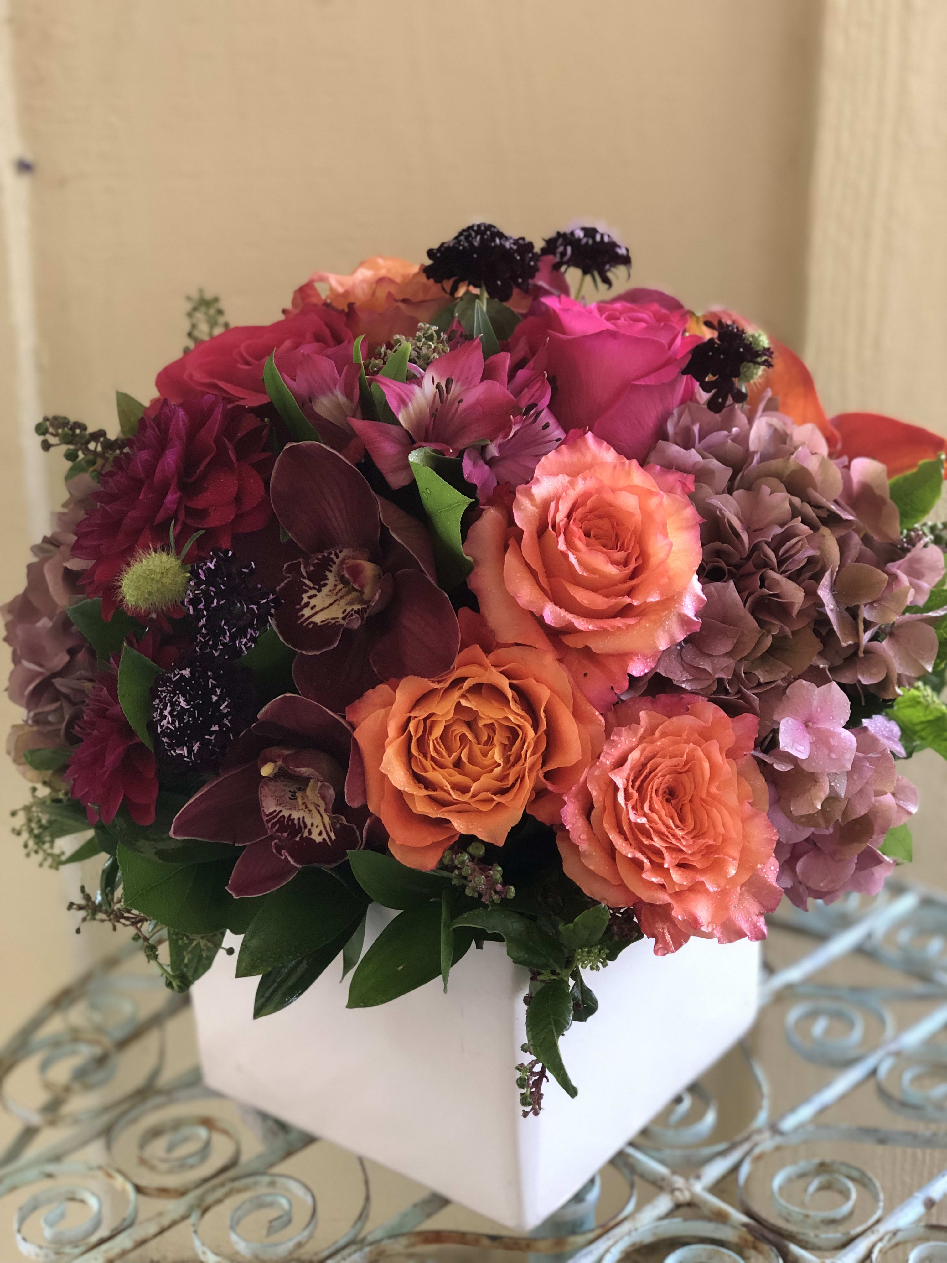 Low cube arrangement of orange roses, pink blooms, and burgundy orchids in a white square vase